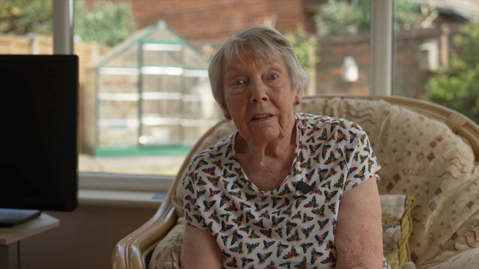 Shiela is sitting on a beige armchair, wearing a white top covered in colourful birds. She has short white hair and blue eyes.