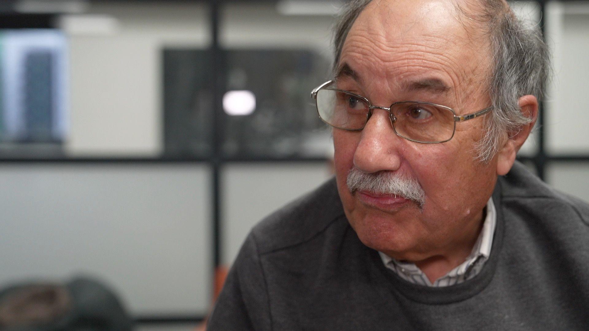 A head and shoulders picture of a man in a grey jumper with a white shirt, wearing glasses. He has a grey moustache and grey hair, and is looking just off-camera.