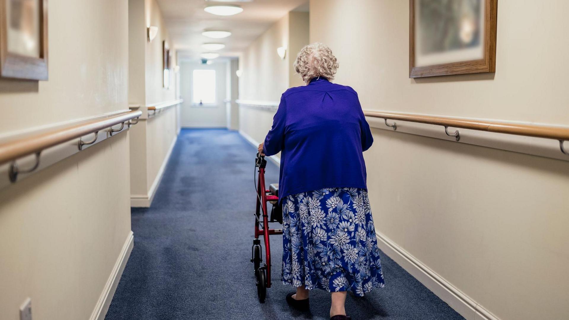 An elderly woman uses a walker to go down an empty corridor with hand rails at either side and pictures on the wall.