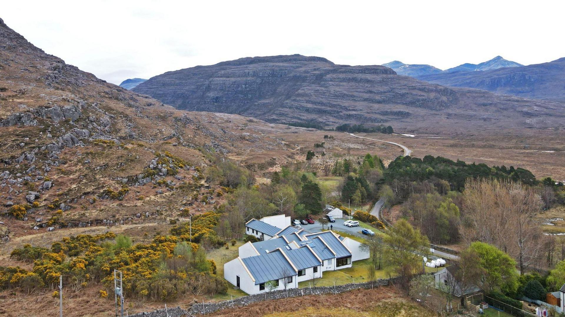 An aerial view of Torridon Youth Hostel. It is a complex made up of low white-walled buildings with grey roofs. The hostel is in a landscape of rugged mountains.
