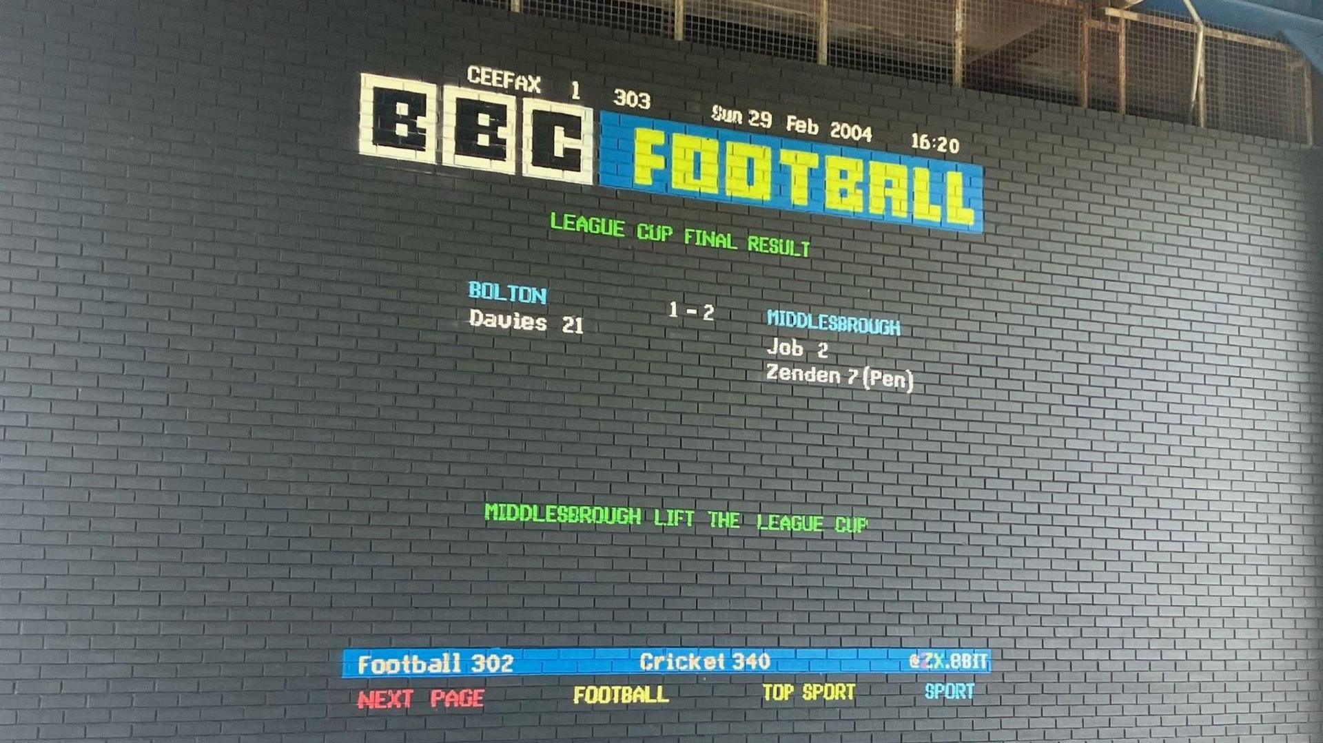 Mural in the style of the former television teletext service Ceefax. Set against a dark grey brick wall it states in red, yellow, green, blue and white letters: BBC Football: League Cup Final Result. Bolton 1 -2 Middlesbrough. Middlesbrough lift the league cup