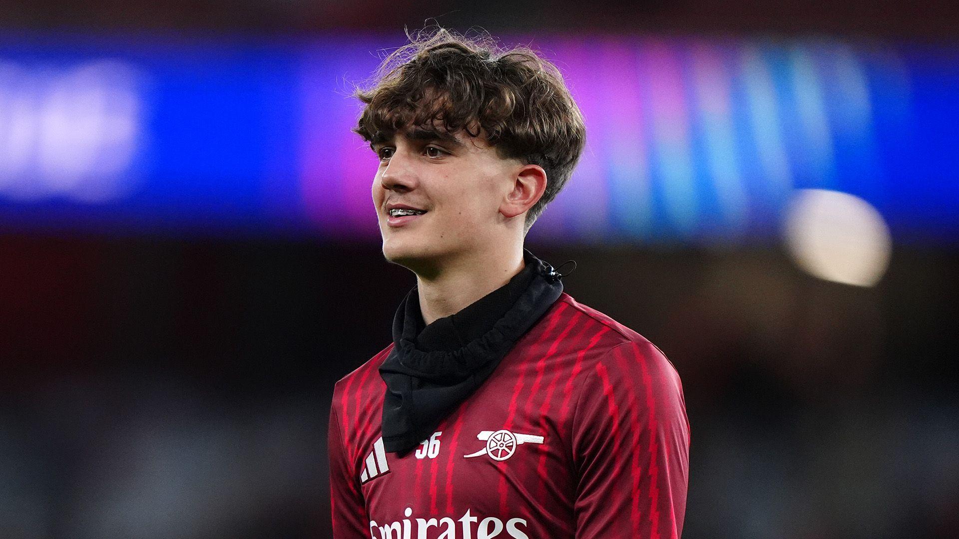 Max Dowman wears a black scarf and his arsenal top. He has short hair which is long-ish and wavy on top. He is smiling and you can see his braces 