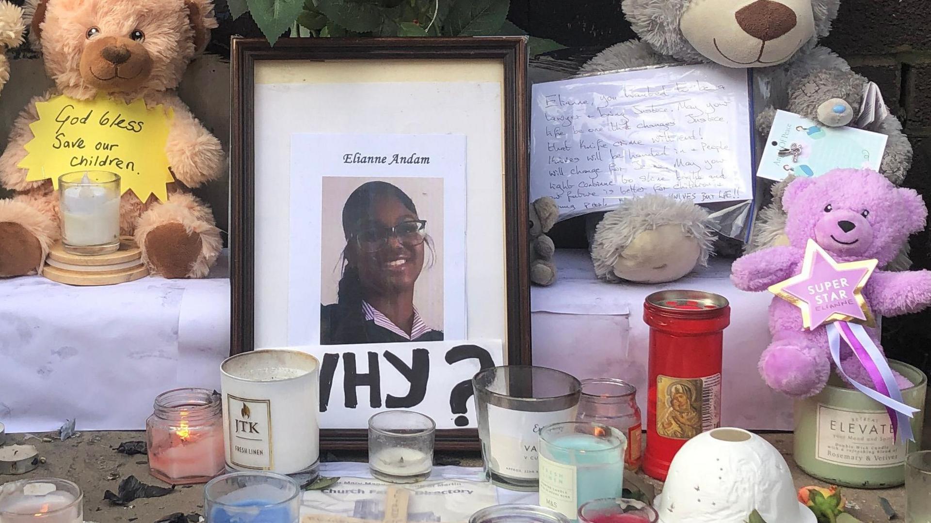 A framed photo of Elianne Andam, smiling, with the word 'Why?' written underneath has been placed on the pavement, alongside flowers, candles and four teddy bears. One bear has a sign that reads 'God bless, save our children'