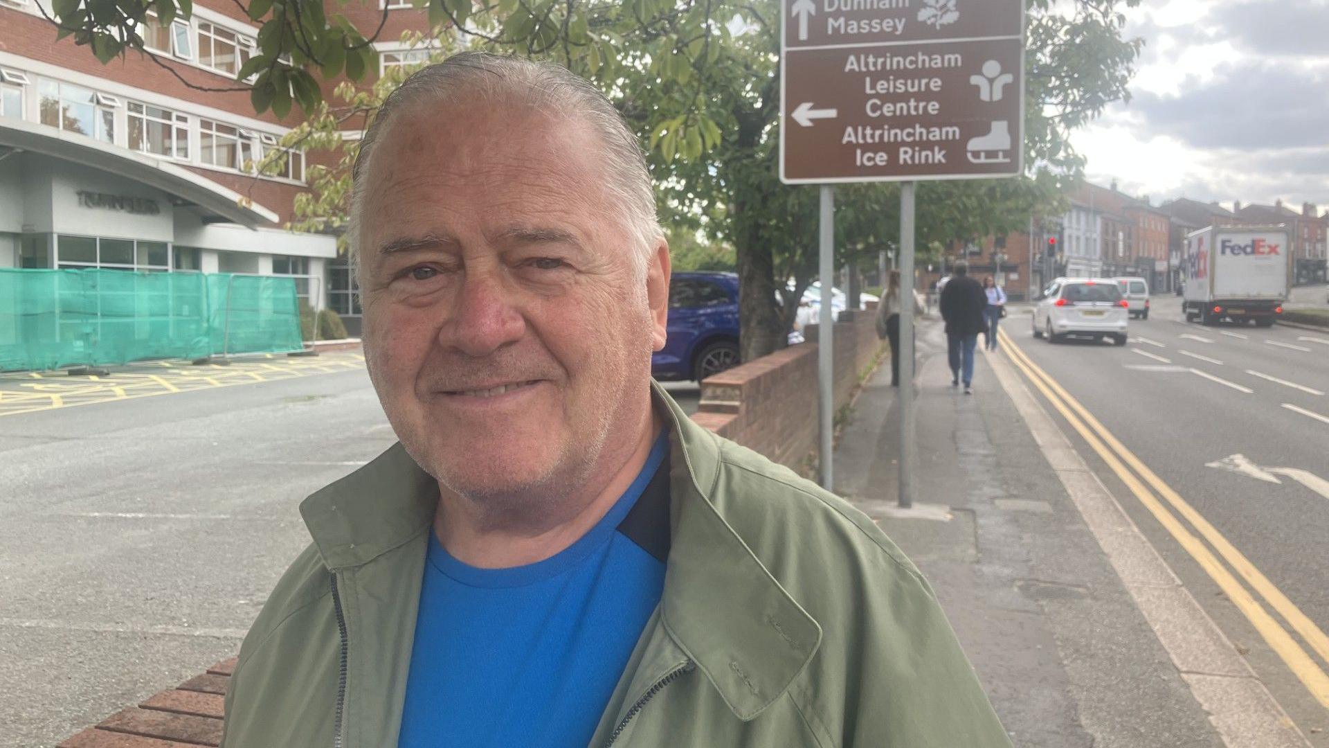Frank McWilliams has thinning grey hair. He is photographed by the side of a road while wearing a green jacket and blue t-shirt.