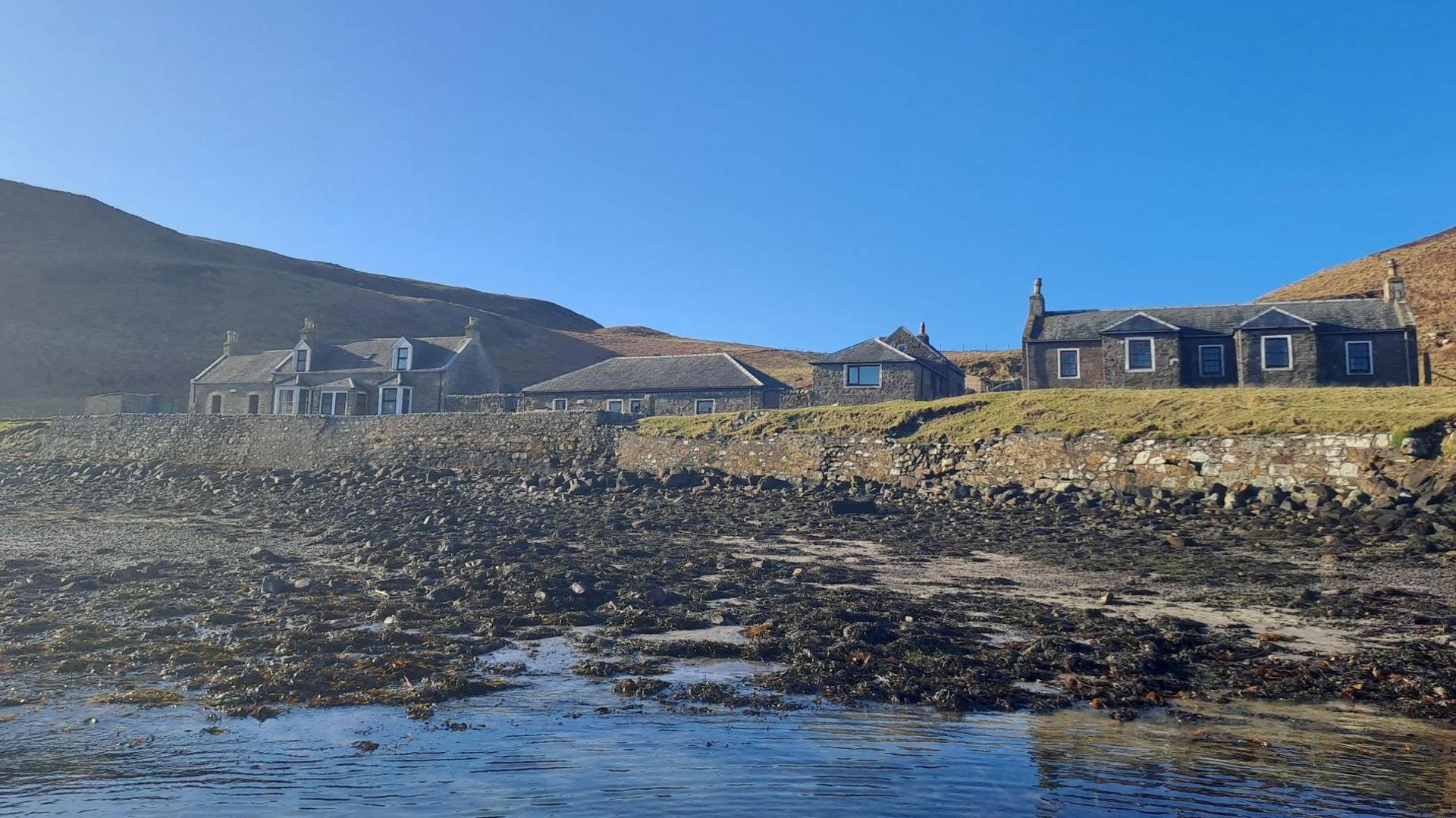 A group of cottages on a shoreline of Sanda. There is a rocky beach and a blue sky above the island.