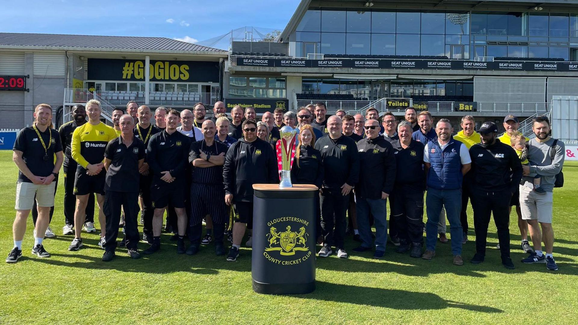 Players and staff of Gloucestershire County Cricket Club stand on the pitch at the county ground in Bristol in a team formation. In front of them is a plinth with the T20 Blast trophy on it. It is a sunny day and the main stand at the stadium is visible behind them