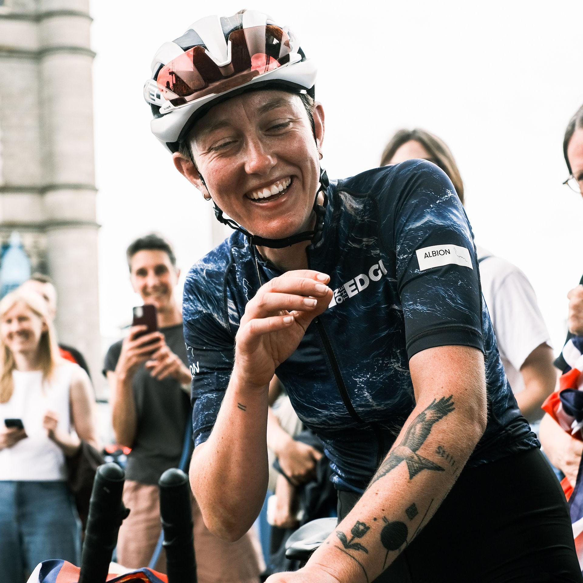 An exhauster woman wearing a red and white cycling helmet and dark blue top, smiles with her eyes almost closed as she sits on a bike. People behind her can be seen smiling and taking pictures with their phones.
