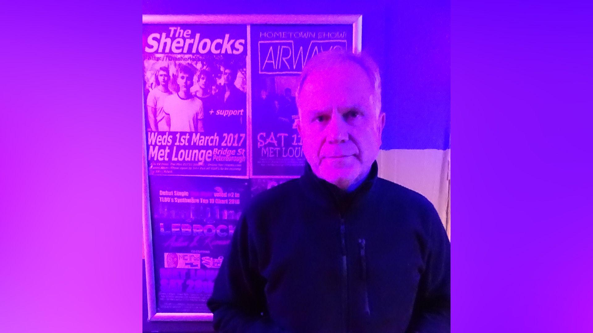 A man with short grey hair is stood in a club under a bright pink light. Behind him are posters advertising the Met Lounge