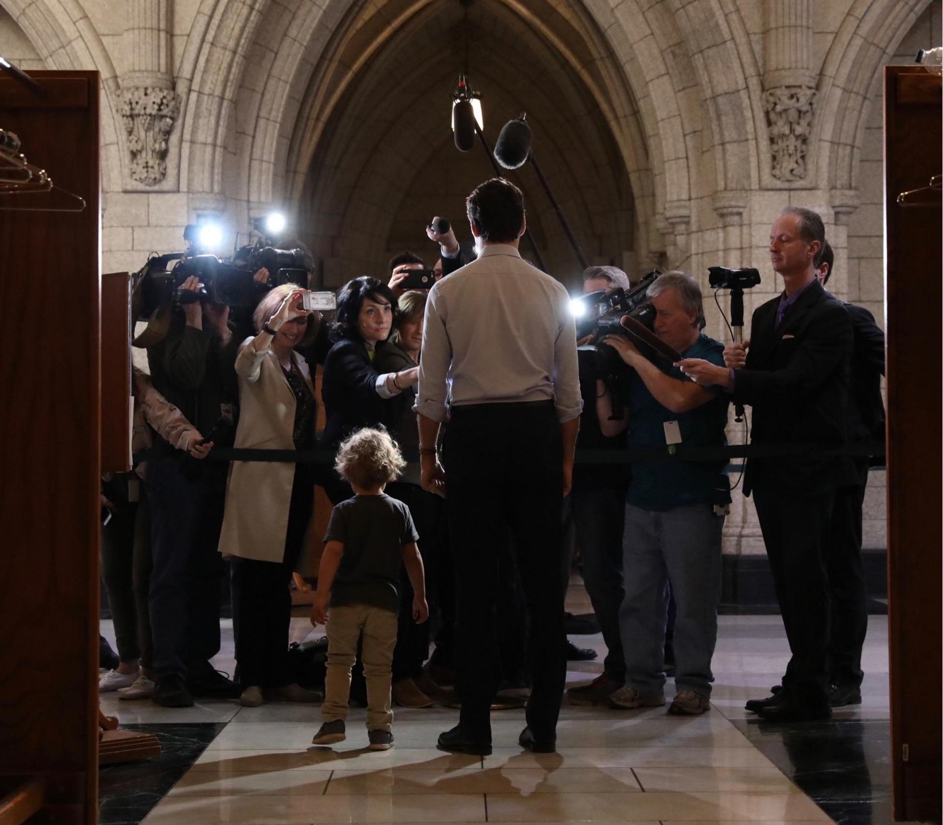 Prime Minister Trudeau gives a statement to the press, flanked by his diminutive offspring, who appears to be distracting some of the reporters.