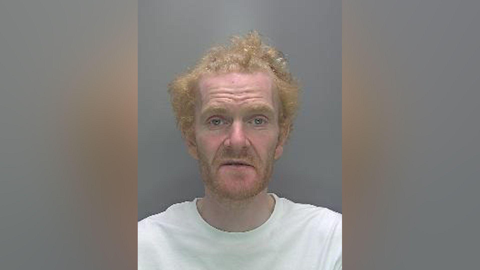 A mugshot of a man with ginger hair and beard wearing a white t-shirt.