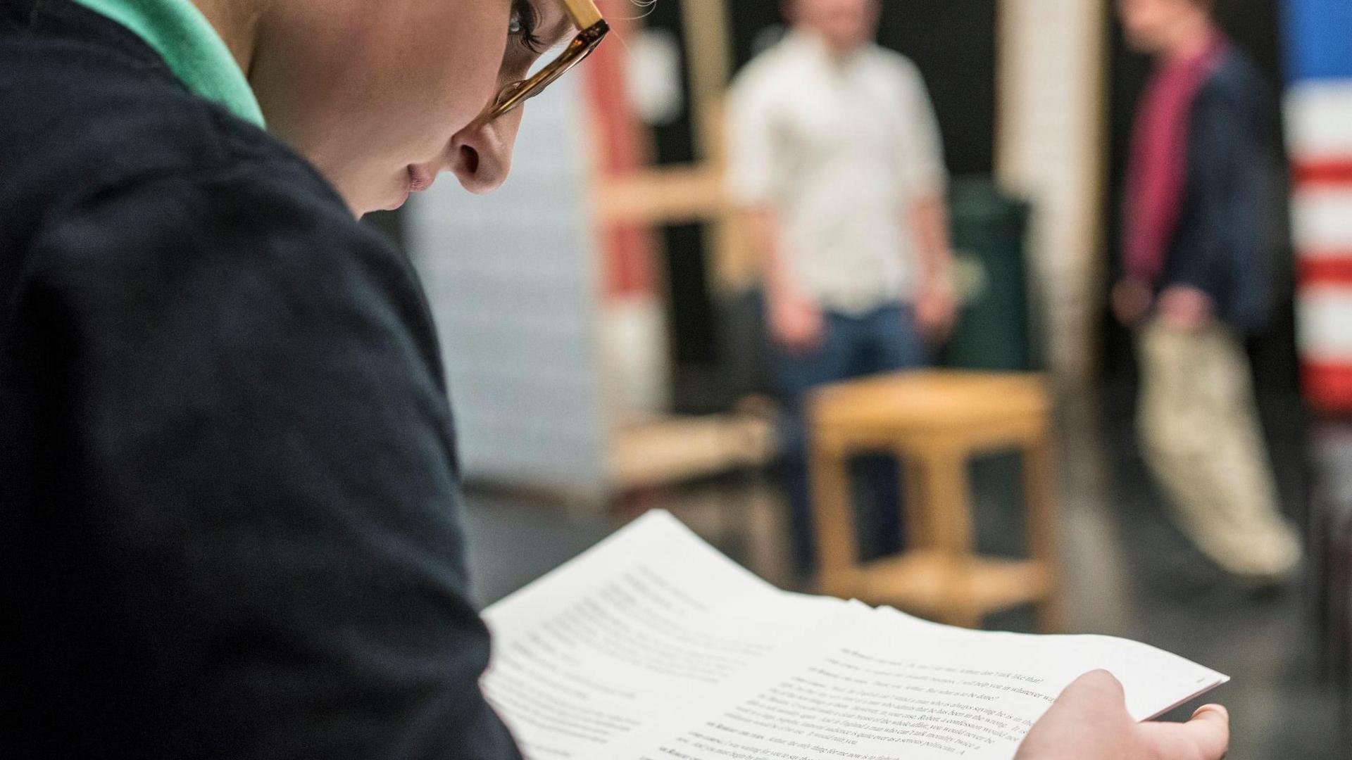 A girl with glasses wearing a black jumper and a green t-shirt holding a script. The view is from over her shoulder and she is looking down at the script. The background is blurred and shows two people standing behind a chair. 