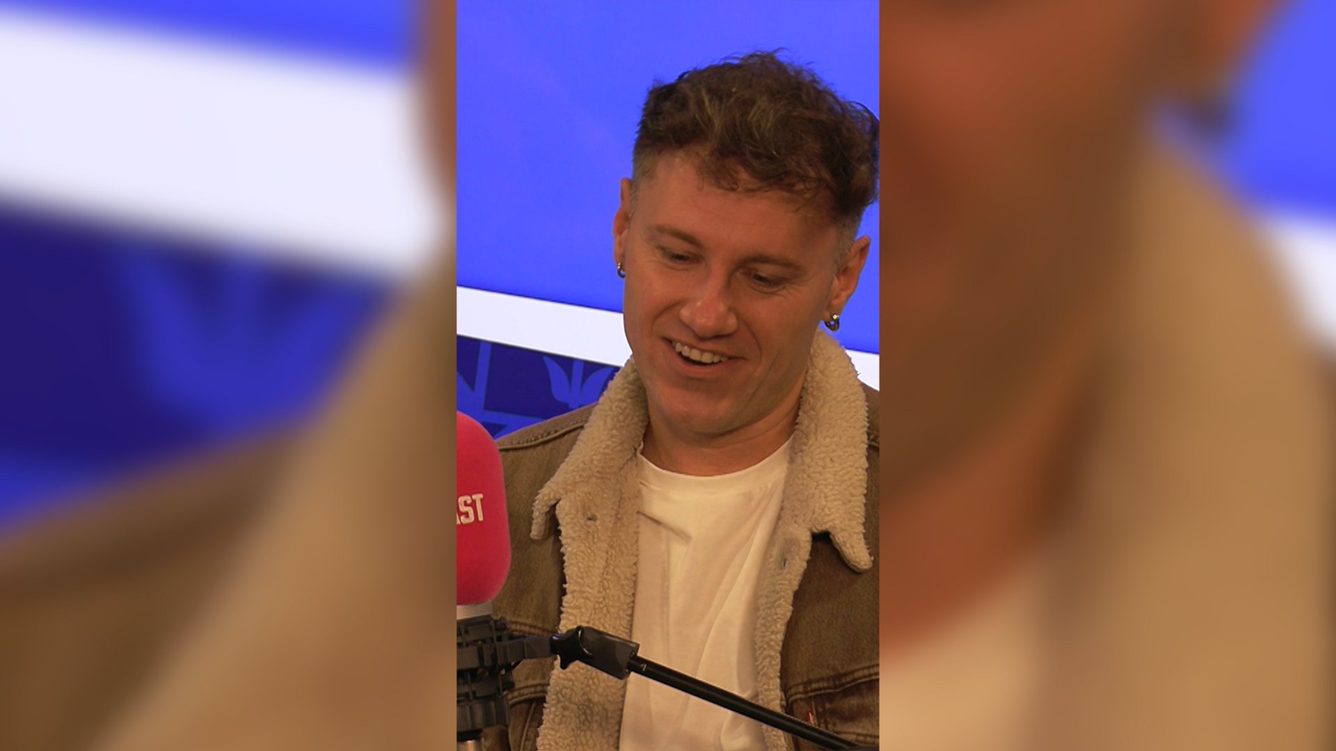 Man with short brown hair speaking in to a pink microphone. He is wearing a grey denim jacket with a fur lining.