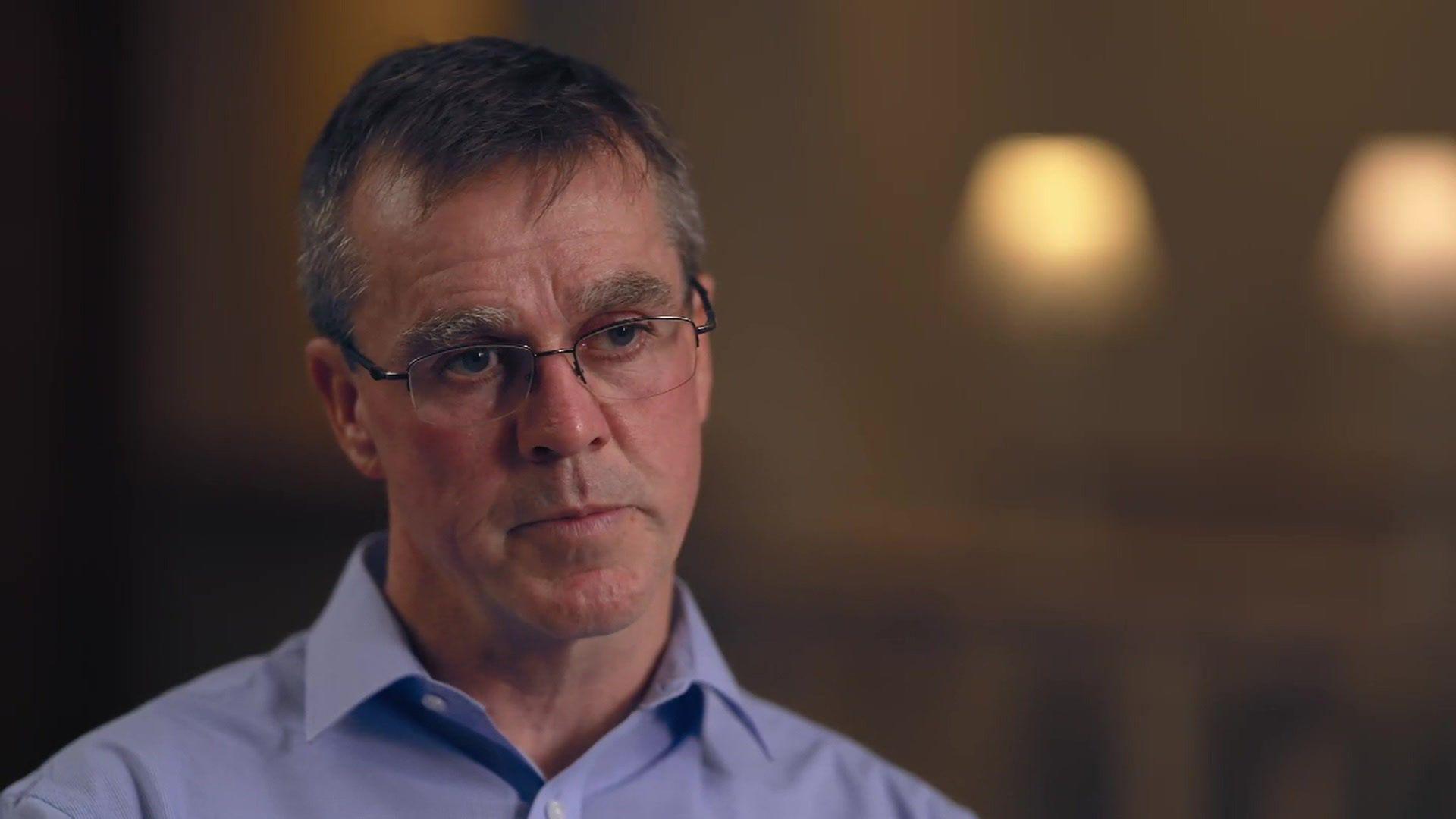 Dr Stephen Brearey, who has short grey hair and glasses and wears a purple shirt, looks into the camera with a serious expression