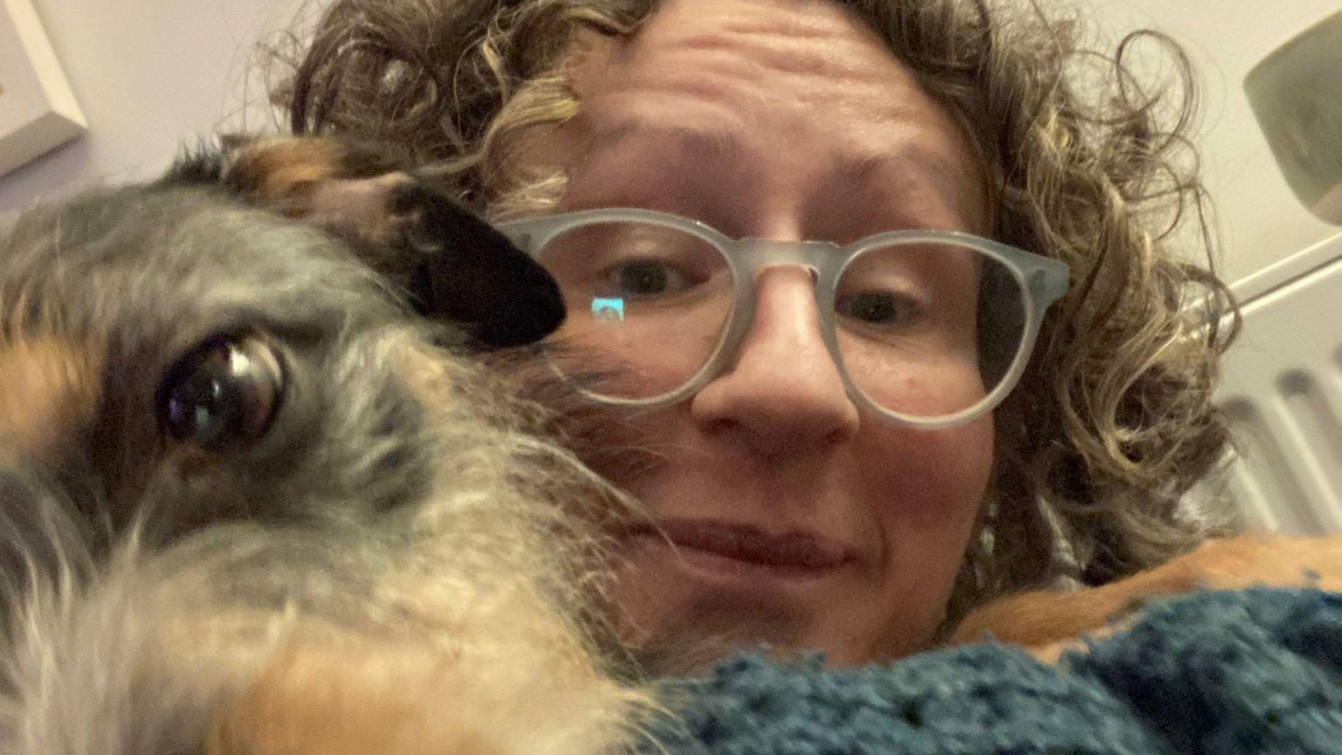 A white woman with curly blonde/brown hair, wearing white round glasses. She is looking directly at the camera. On the left of the photo is her dog's face. It is brown and blonde, with brown eyes.