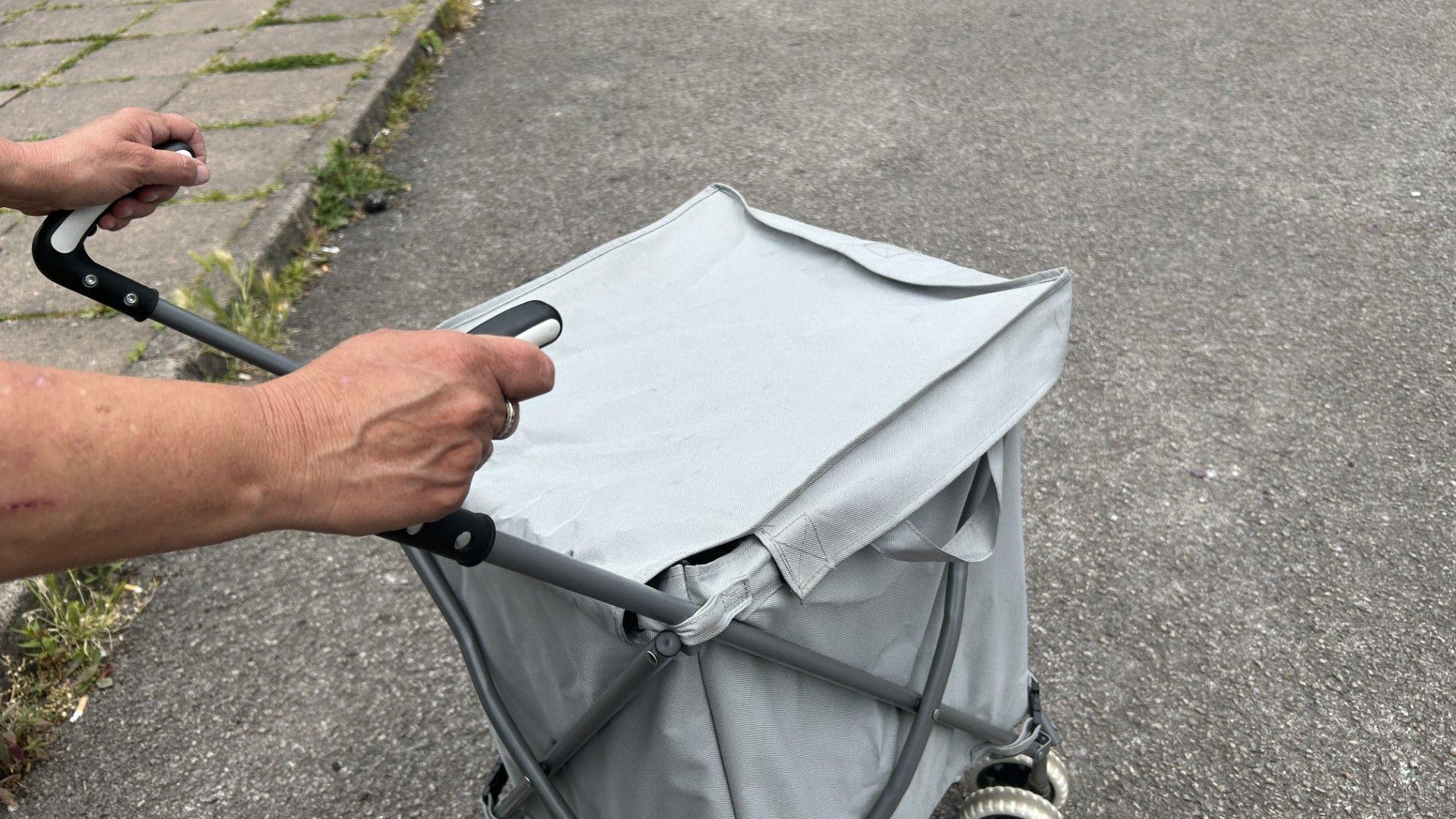 A woman holding a shopping trolley. It is grey. We can see her hands gripping on and her veins. There is a concrete floor in the background.