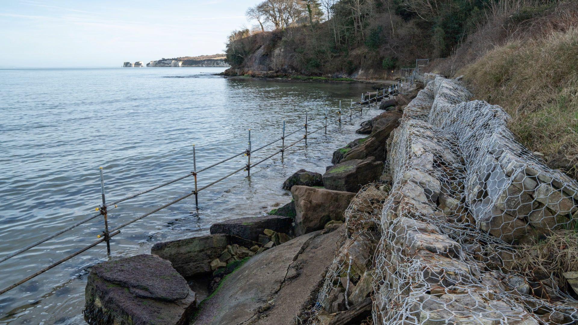 A view of some of the stones wrapped in metal wire along the seafront as a defence against the water. There is also a short metal scaffolding fence in front of some exposed rocks in the water. In the distance some cliffs with green grass on the top can be seen.