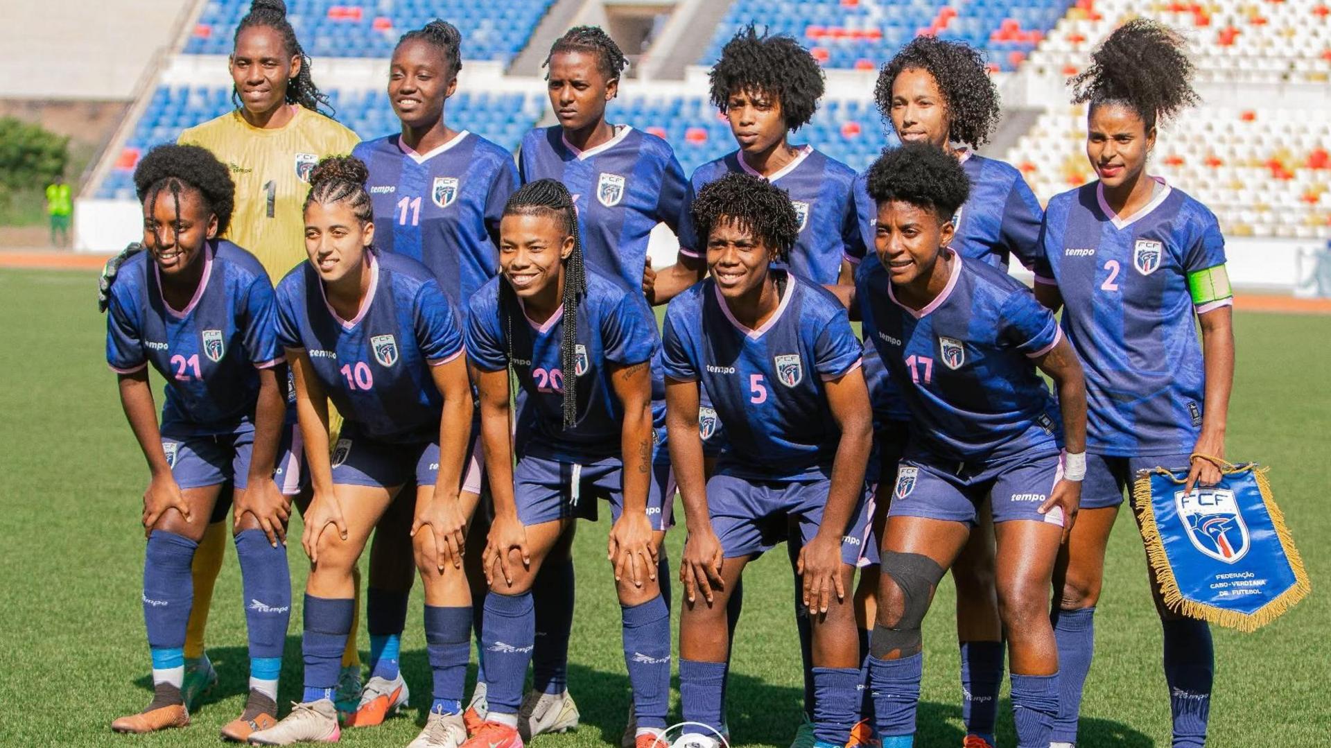 Cape Verde's women's footballers pose for a team photo before a game on a sunny day. Their goalkeeper, positioned far left of the back row, wears a golden jersey while the 10 outfield players are wearing an all-blue strip. The six players in the back row stand with arms around either other's waists, with the captain on the far right holding a national team pennant. The five players in the front row are standing but leaning forward with both hands on their knees