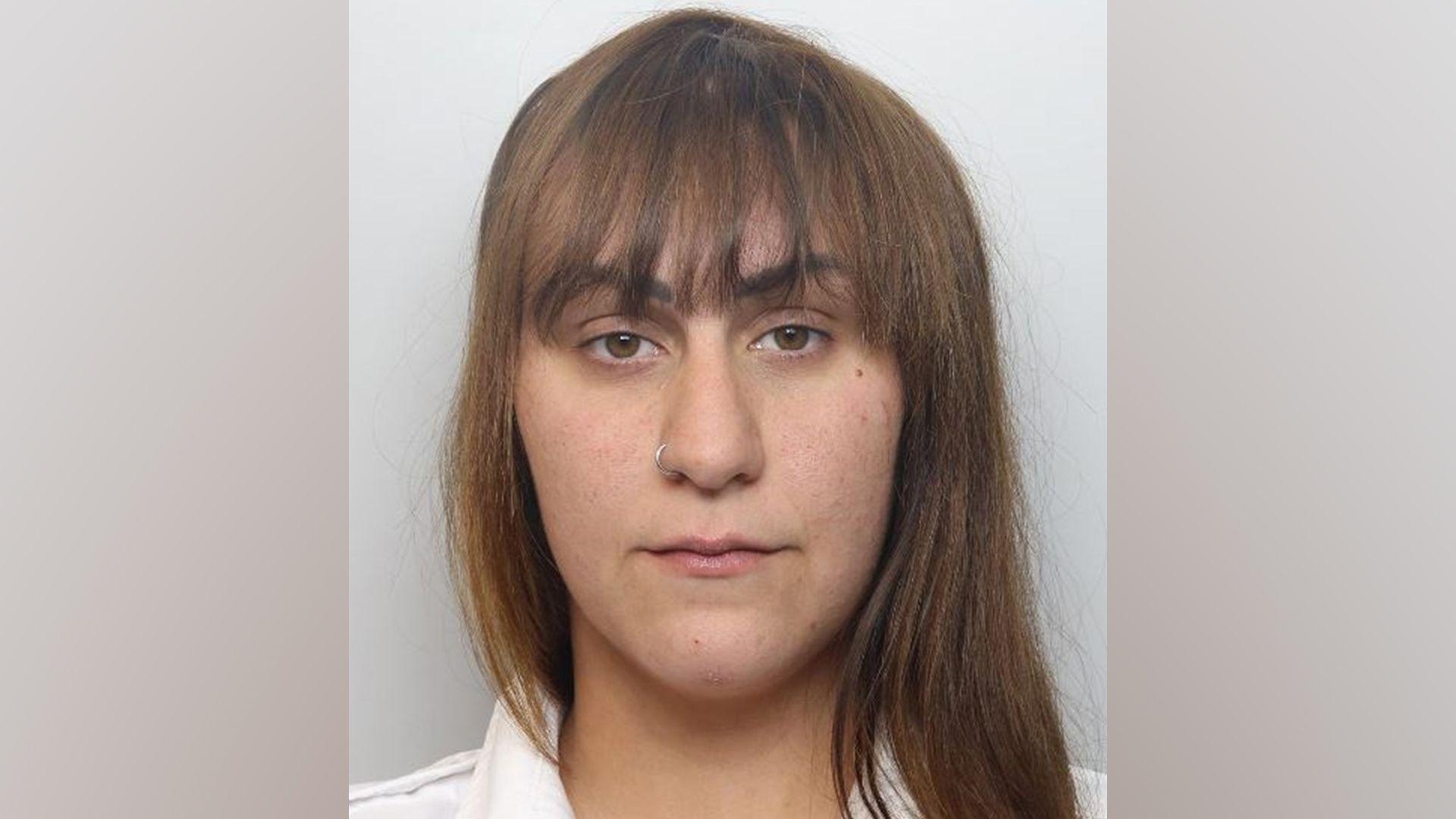 A mugshot photo of a woman with long brown hair stood against a grey background. She has a nose ring piercing.