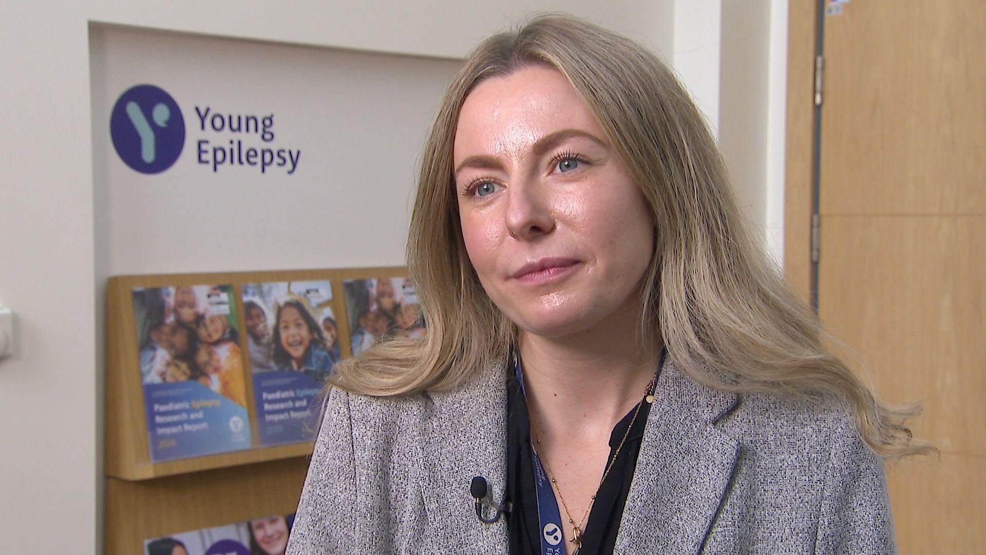Lara Carr wearing a grey jacket and black blouse with Young Epilepsy brochures on a shelf in the background 