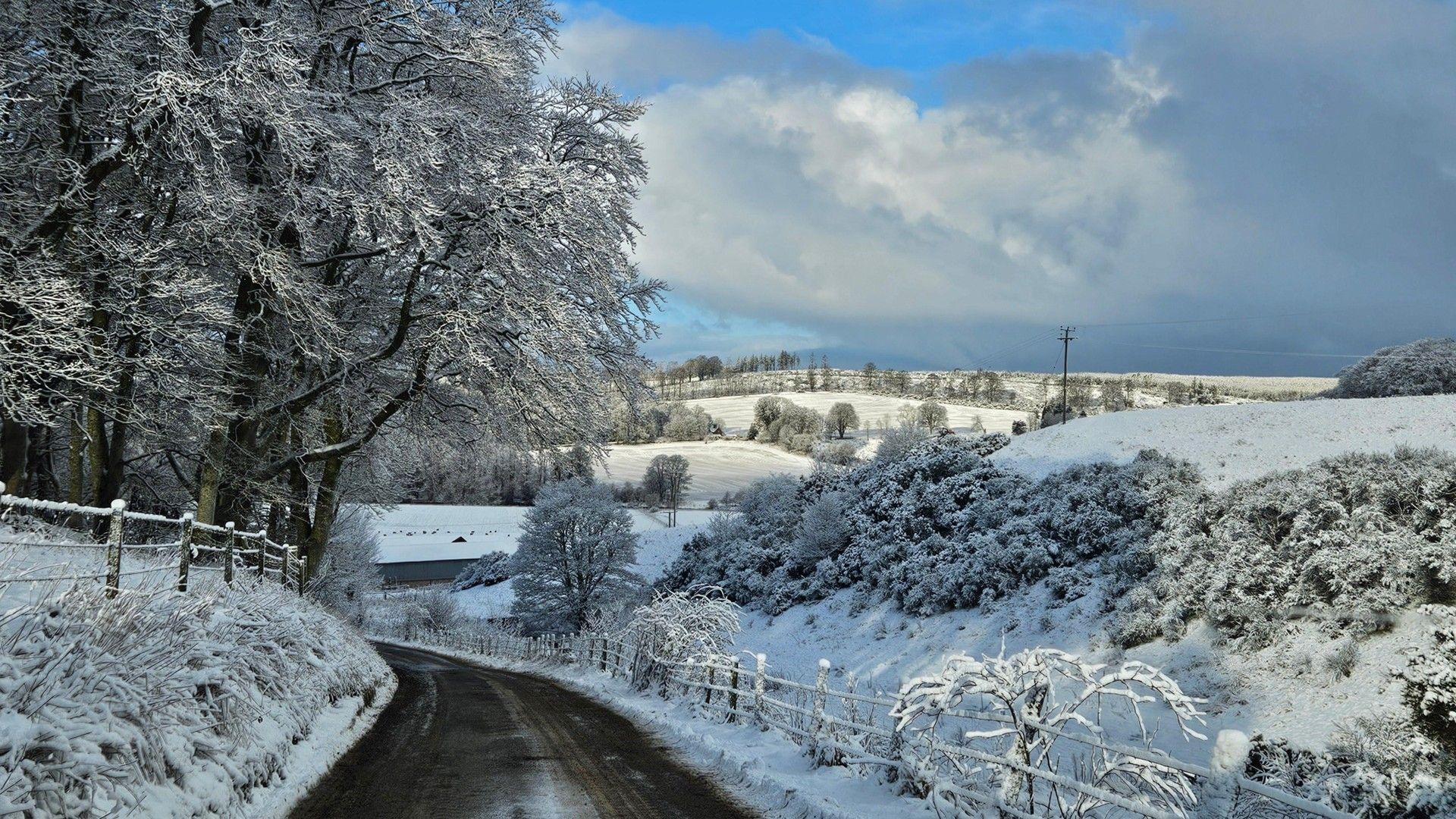 Snow has been cleared from the road but there's a good covering of snow in the fields and on the trees with side. Broken cloud with some blue sky overhead.