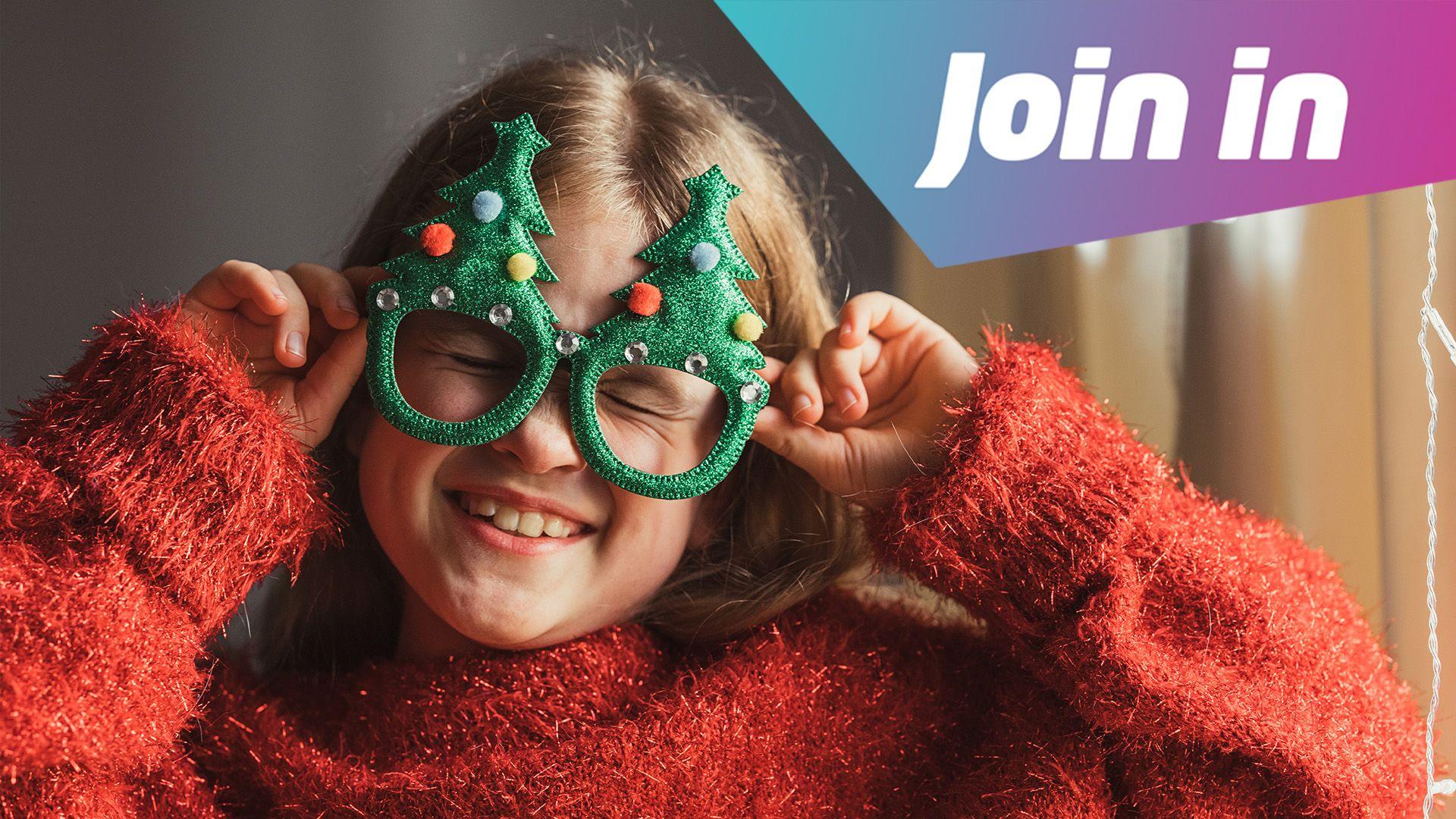 A girl wearing a red fluffy jumper and holding Christmas glasses on her face. They are shaped like Christmas trees and are green with added coloured pom poms. She is smiling and screwing her eyes up