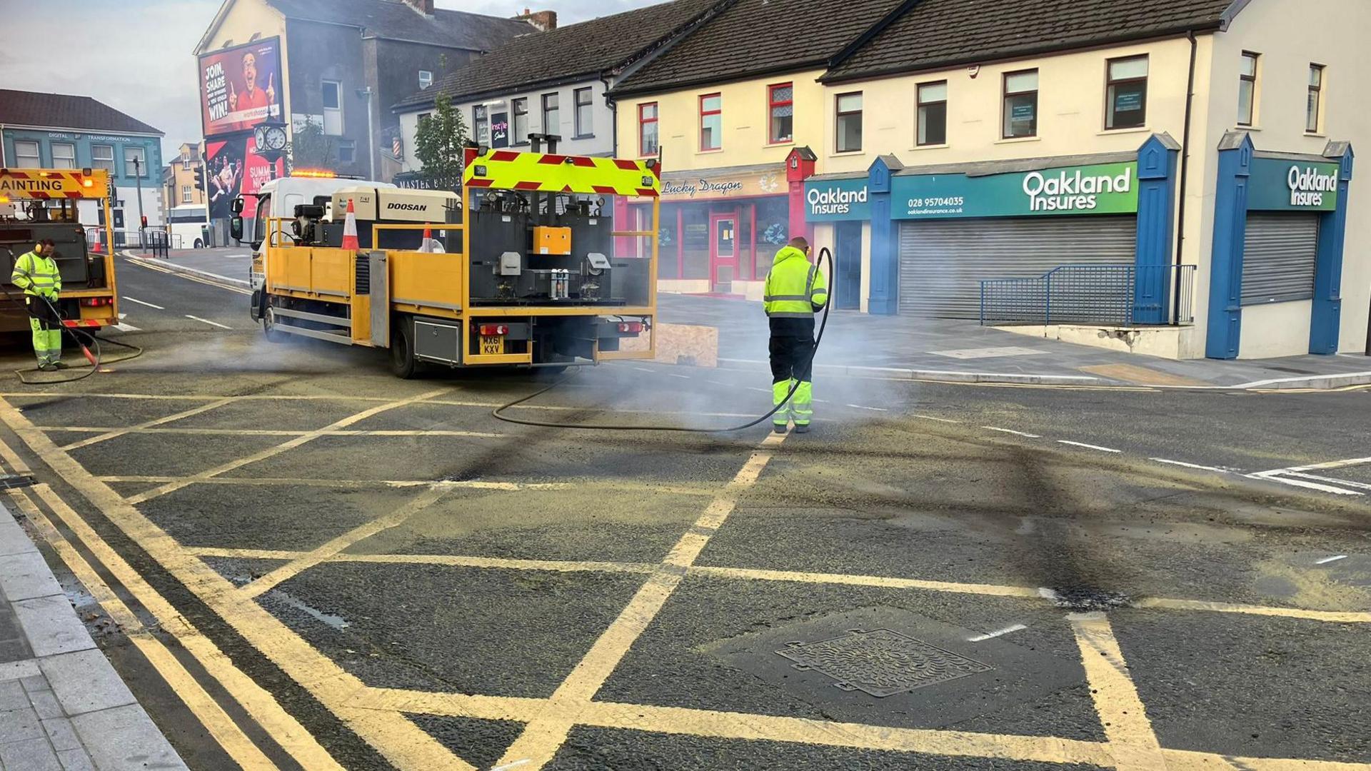 The picture shows work men blasting off some of the yellow paint in a large box junction in Maghera. They are dressed in yellow high-viz clothing and two yellow lorries are also in the picture