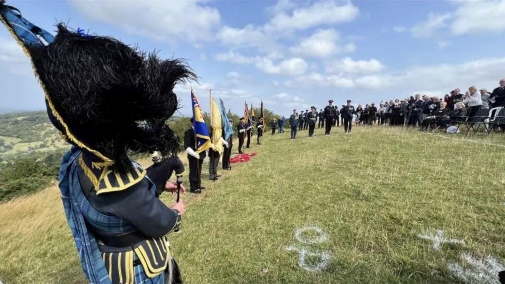 A commemorative service being held on Cleeve Hill. People are standing holding military flags while a bagpiper plays in the foreground