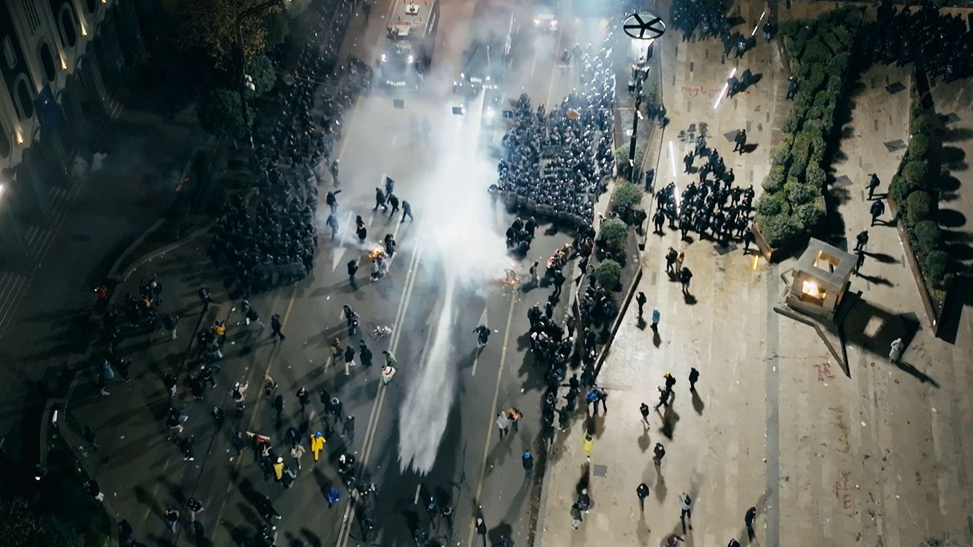 Crowds scatter as water is sprayed out from trucks in the Georgian capital - there is a large plume of water and widely dispersed mist. It is dark and the shot is taken from above.
