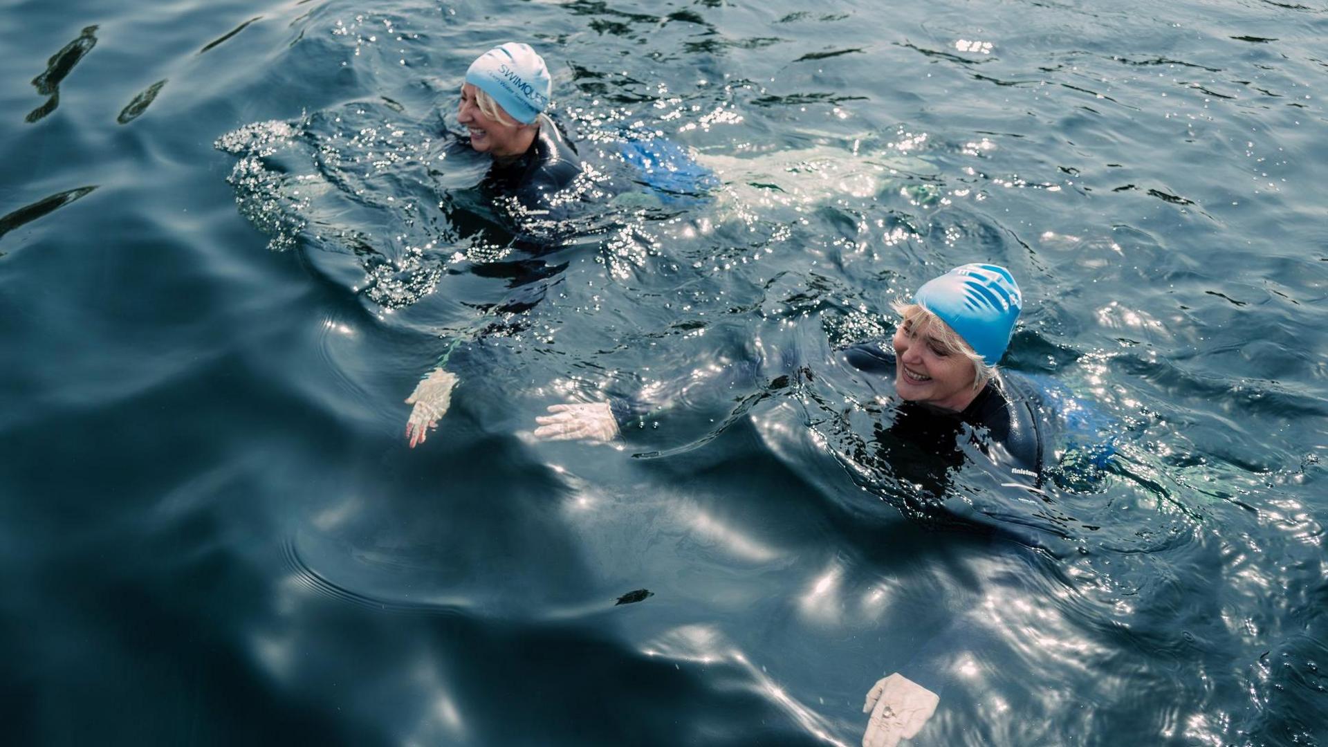 Actors Miranda Richardson and Maggie Service are swimming alongside each other. They are wearing light blue swimming hats.