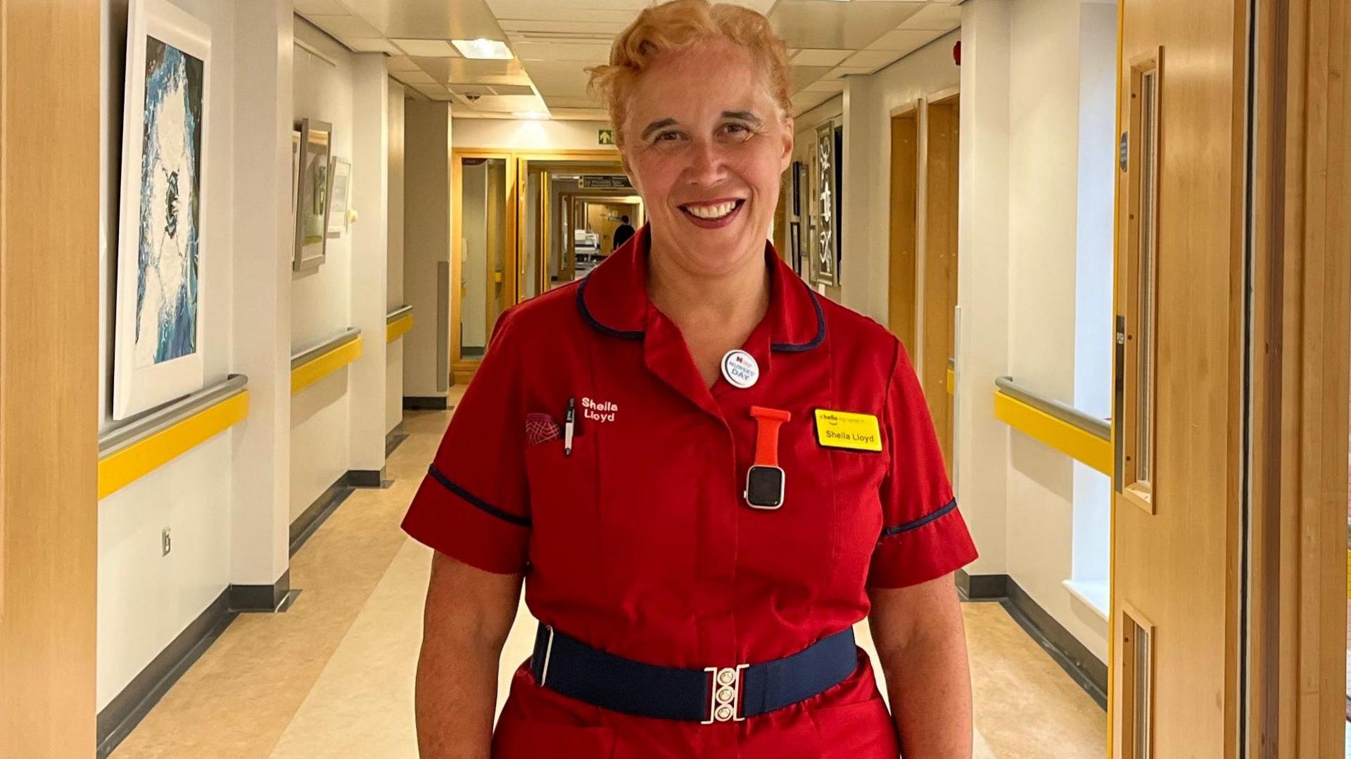 Sheila Lloyd, a woman with blonde hair in a bun and a bright smile, she is wearing a red nursing dress and is standing in the hospital corridor.