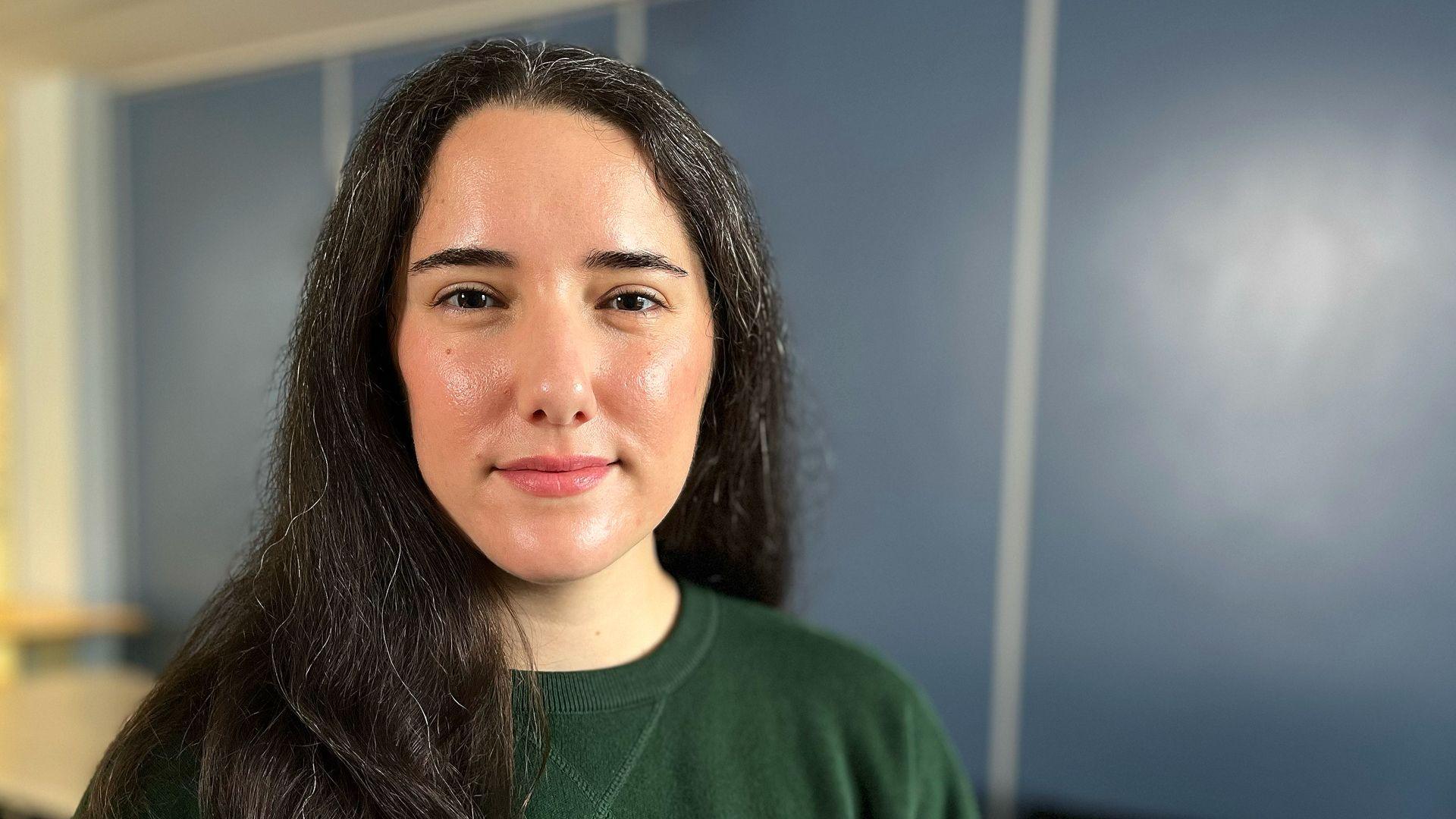 Stephanie Metzger wearing a dark green sweatshirt with long, dark hair, standing indoors in front of a blue wall with vertical panel lines. The background includes a blurred light-colored surface on the left side, suggesting a table or counter.