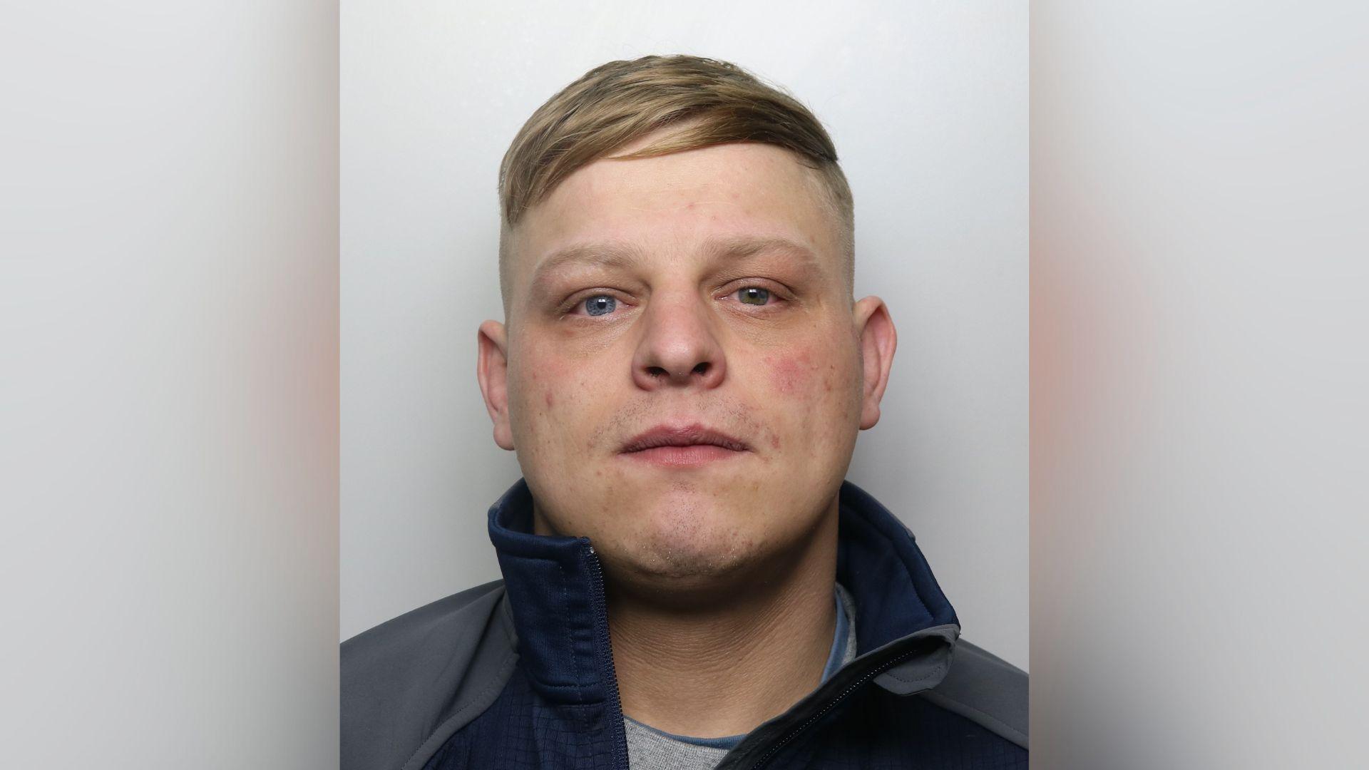 A police mugshot of a Liam Woods, 25. He is blond, with a wide face. He has blue eyes and some pink blemishes on his cheeks. He is looking directly at the camera with a fairly neutral expression.