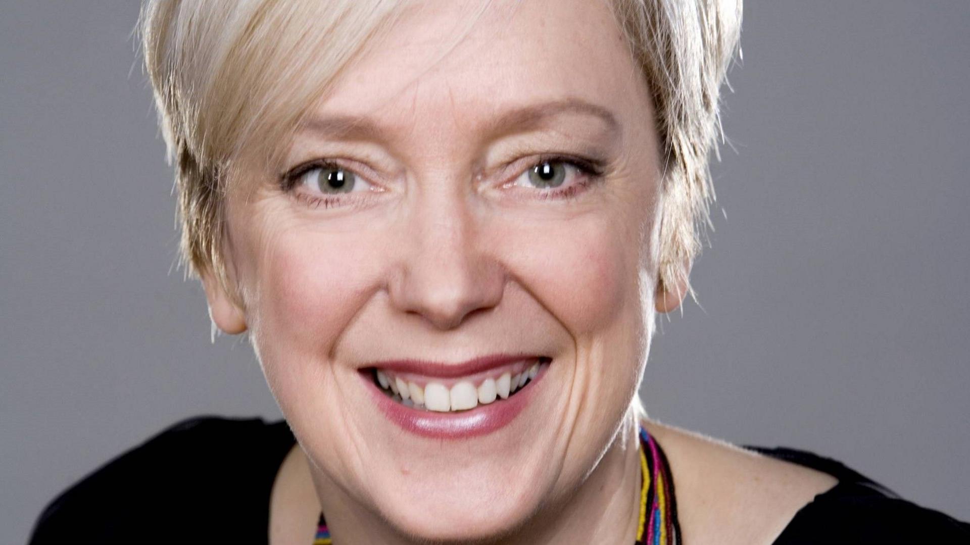 Janice Forsyth, a woman with cropped blonde hair smiles at the camera. She is wearing pink lipstick and a black top.