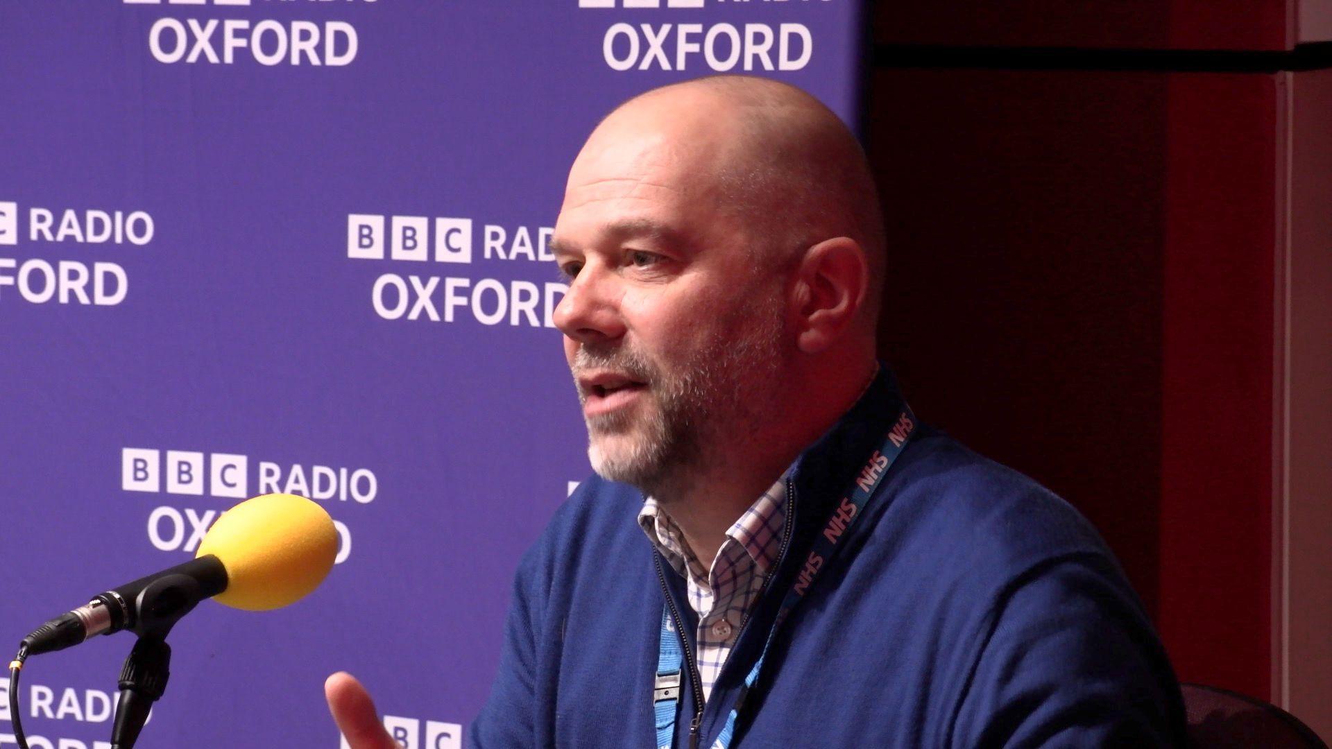 Dan Leveson, Place Director for Oxfordshire at Buckinghamshire, Oxfordshire and Berkshire West Integrated Care Board (BOB ICB) speaking on BBC Radio Oxford into a yellow microphone, in front of a BBC Radio Oxford branded banner.