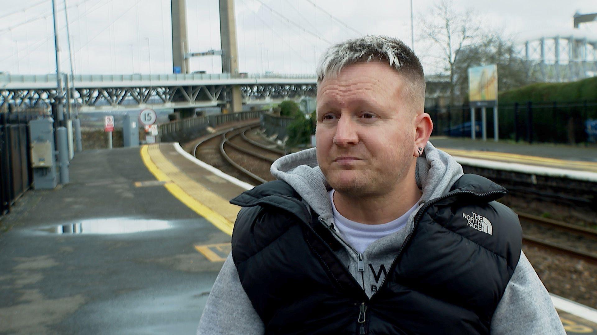 Scott has short hair and is wearing a grey hoodie under a black body warmer. He is pictured in front of a railway track at Saltash station