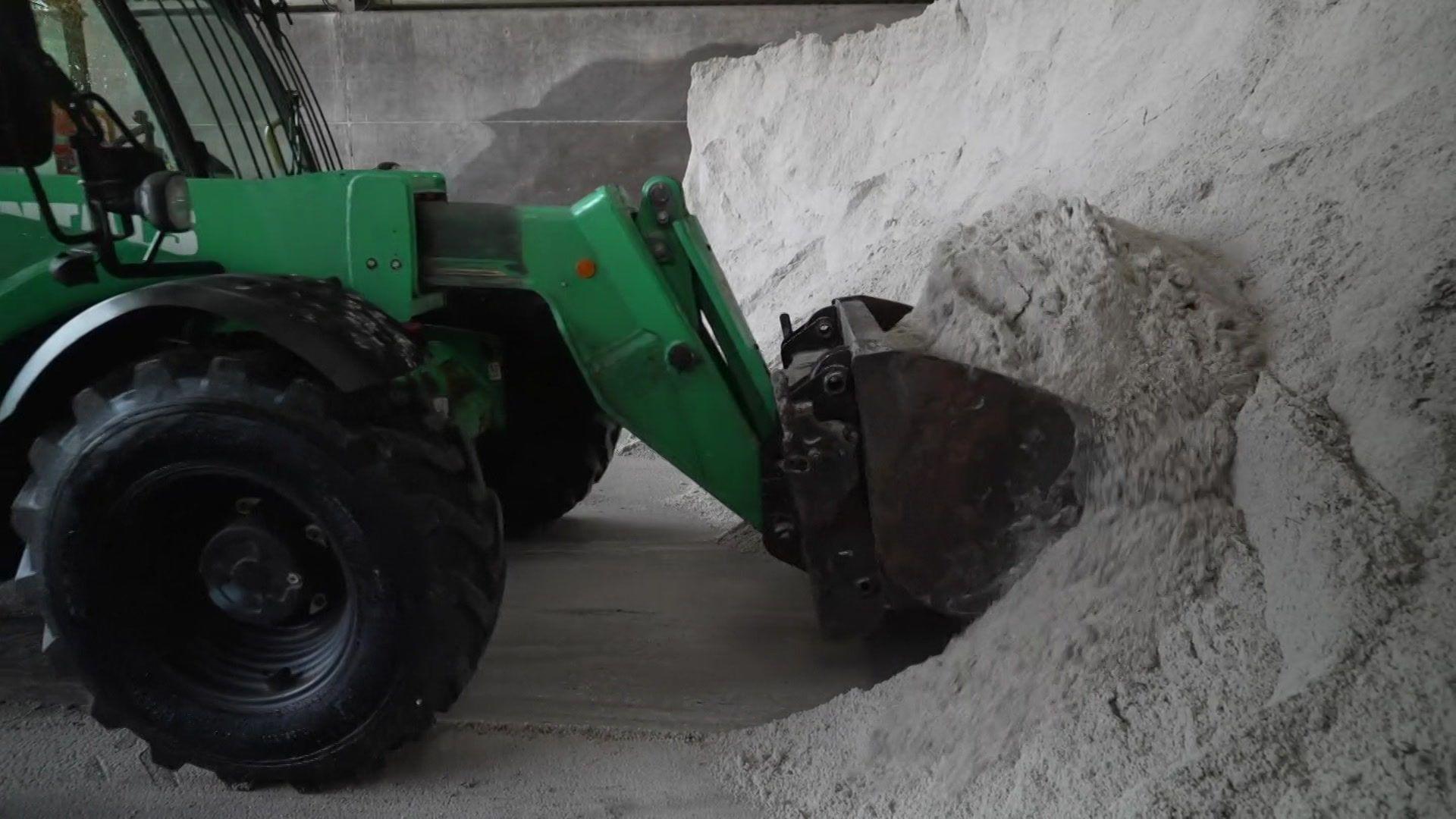 A green tractor with a scoop on the front shovelling a pile of grey gritting salt.