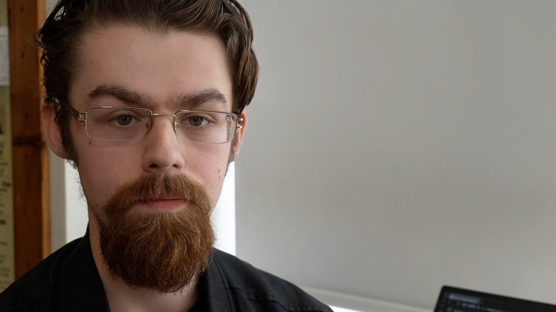 Reece Stone looks expressionless at the camera. He has brown hair with a red beard. He wears a black top and small glasses.
