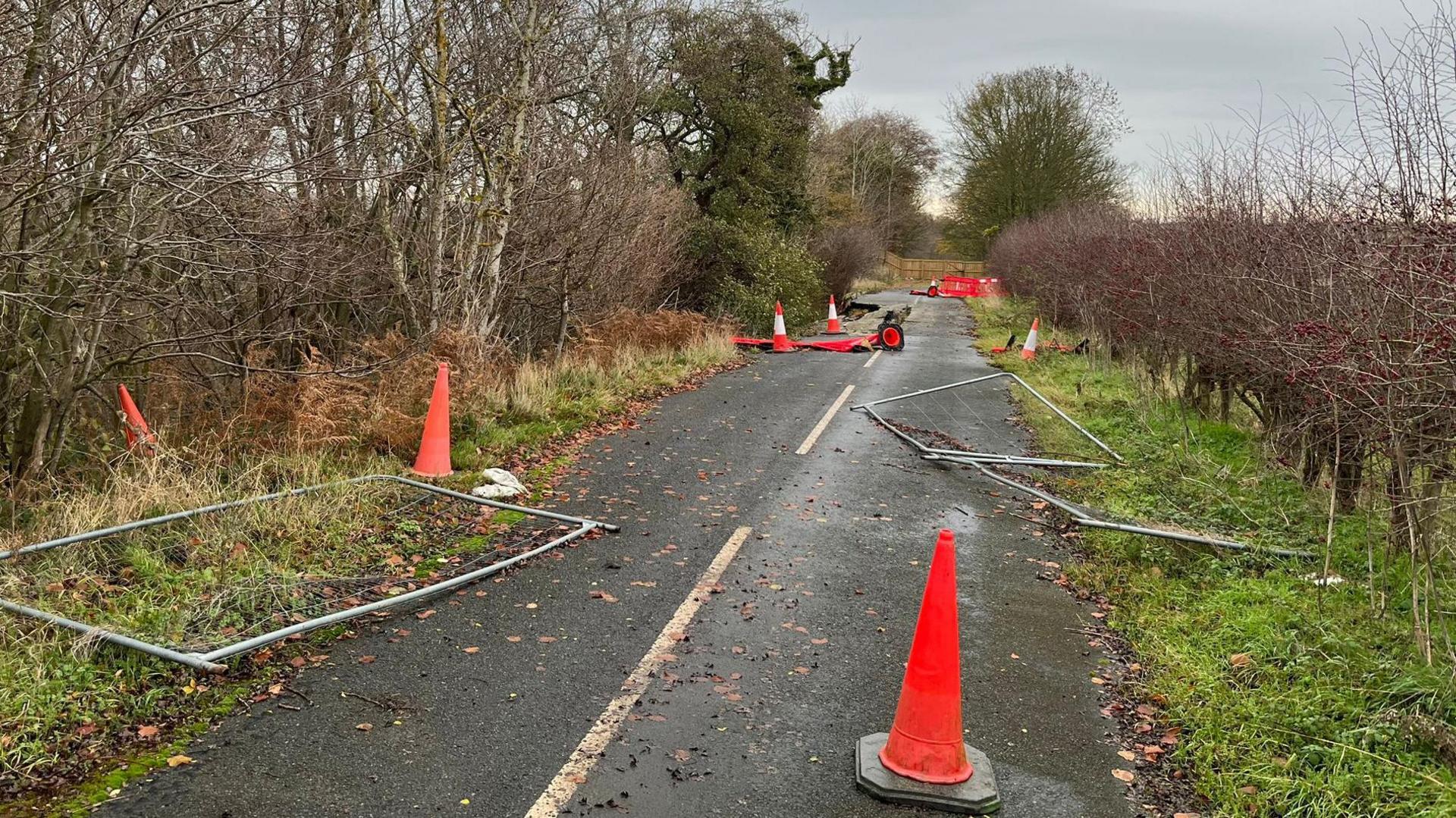 A fenced off part of road with metal fences lying on the ground with traffic cones dotted around. Further up the road part of it has caved in and more barriers can be seen fencing off pieces of tarmac.