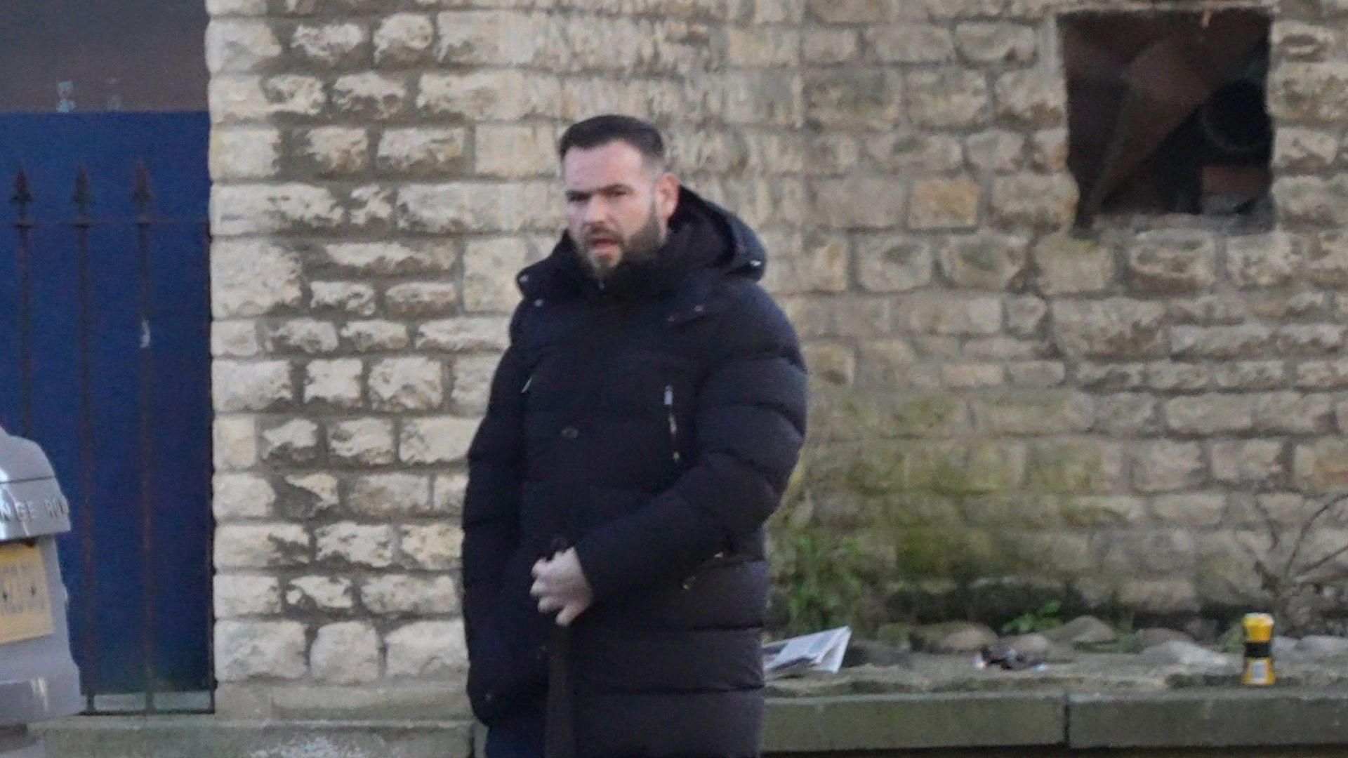 Fred Doe, who has short black hair and beard, looks at the camera. He is dressed in a black puffer coat and is standing outside by a stone wall.