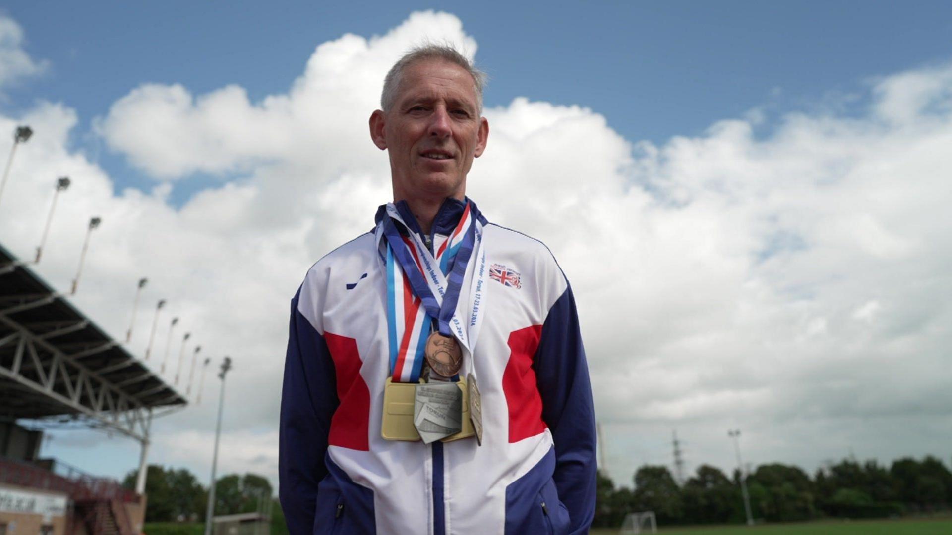 Darren Scott, with a number of gold, silver and bronze medals around his neck, stands looking into the camera against a backdrop of an athletics stadium