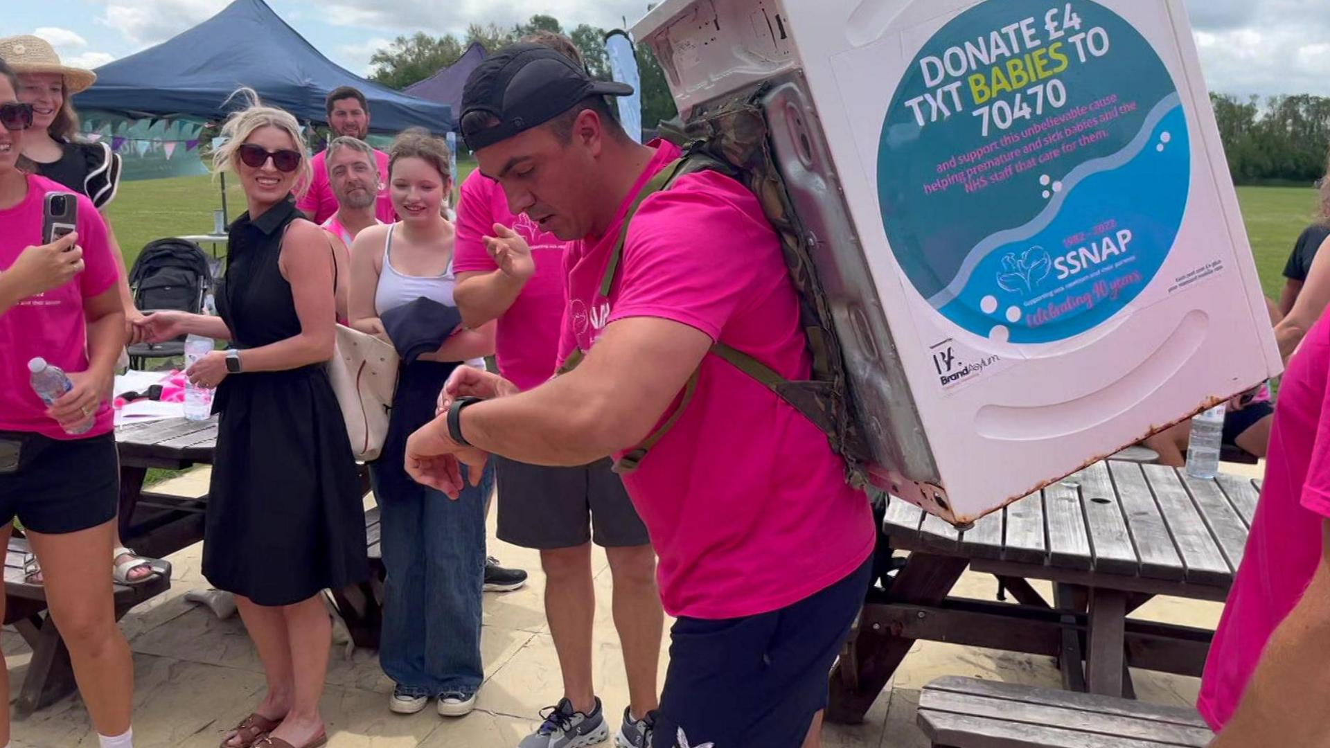 A man checks his watch before starting his running challenge. He wears a pink shirt with the SSNAP logo, blue shorts, and a blue cap back to front. He has a washing machine on his back.