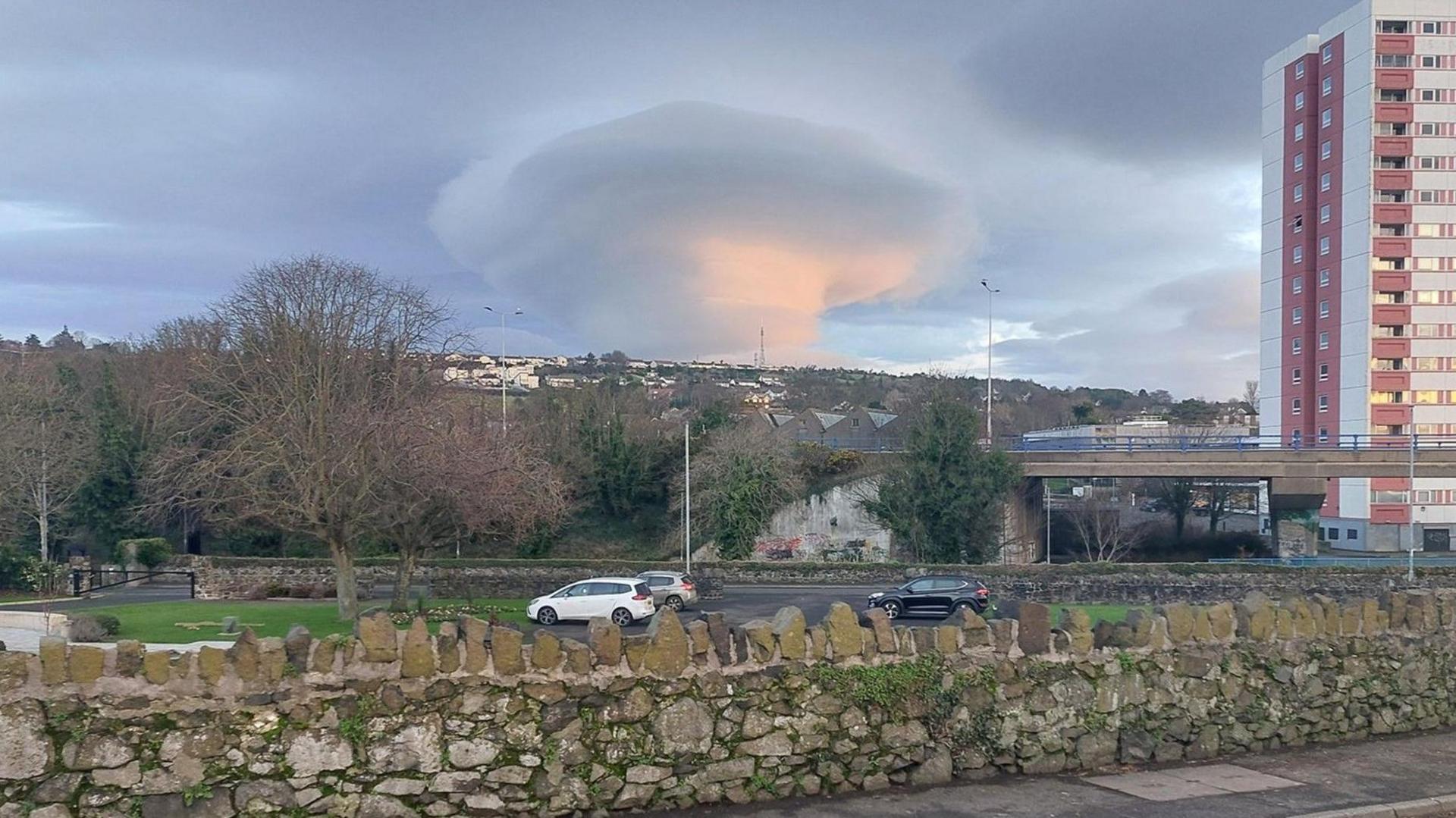 A stone wall, with the view of a town in the distance and a high rise building to the right. The sky has a large cone shaped cloud.