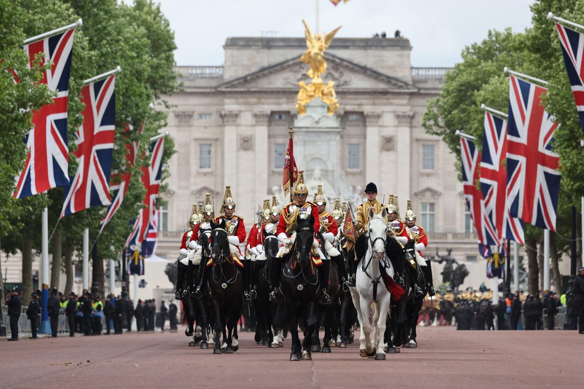 The procession of military personnel riding horses parade down the Mall with union jacks and green foliage lining the street and Buckingham Palace in the background.