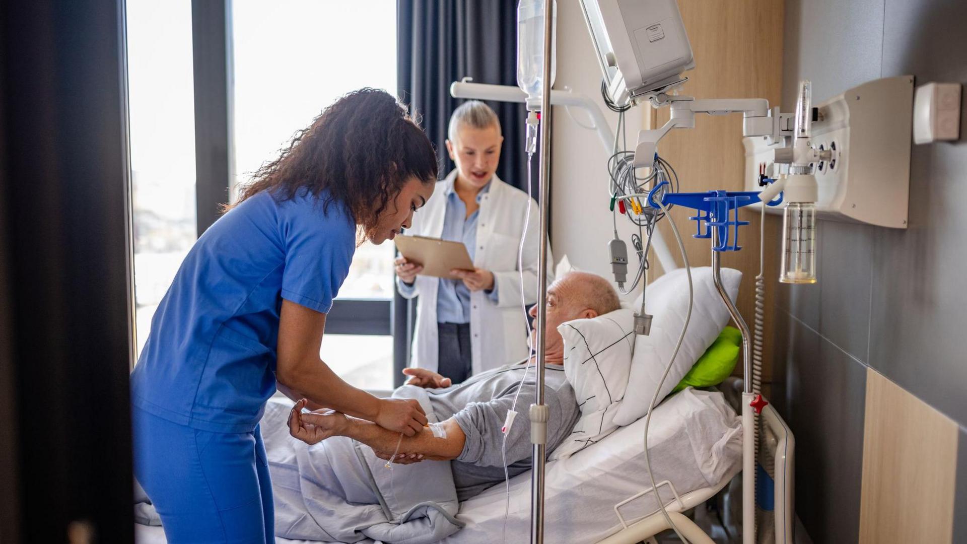 A hospital patient lying in a bed is being tended to by a nurse wearing a blue uniform, and a doctor in a white coat is standing behind the bed, in front of a window