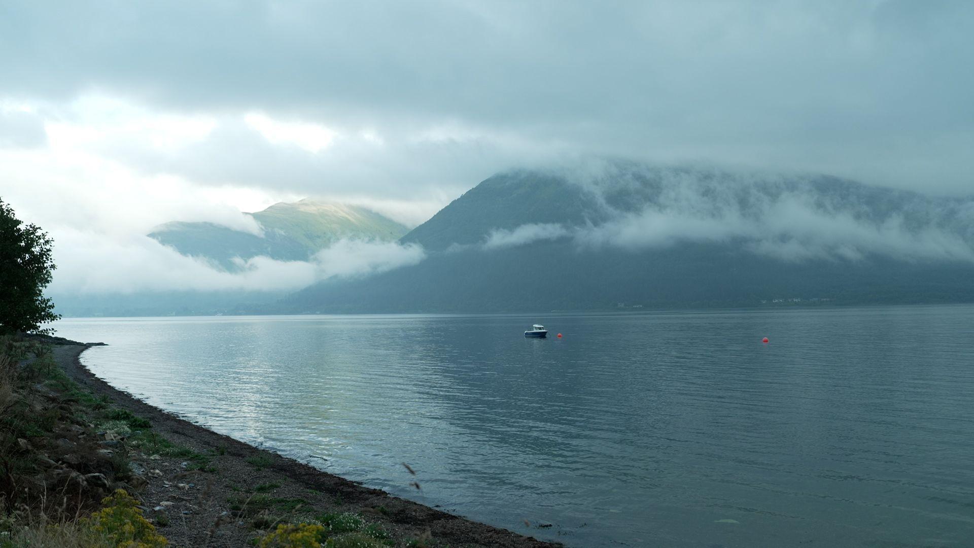 A stunning landscape of a Scottish loch, calm and empty, save a lone small fishing boat. Mountains frame the background and mist weaves its way up and down the outline of the hills