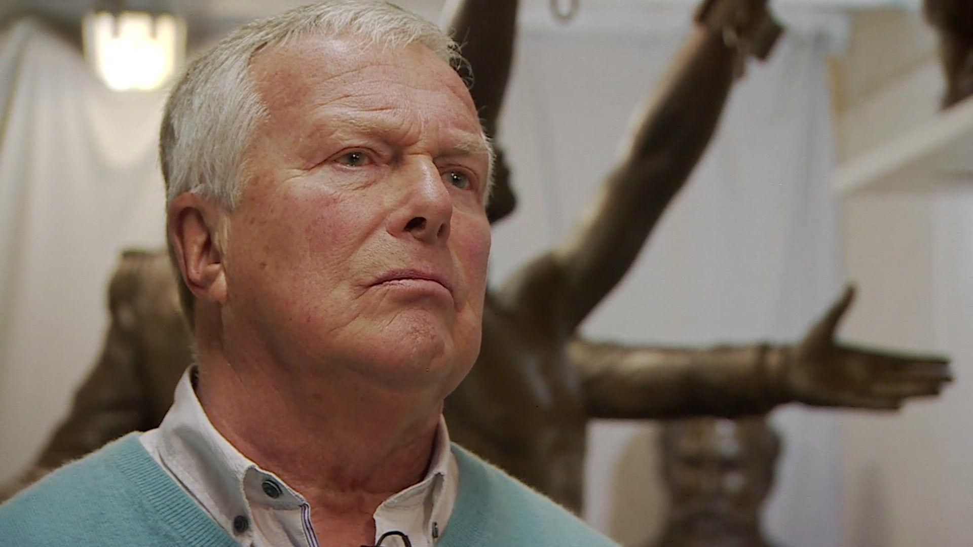 Colin Kemp standing in a workshop in front of a sculpture. He is wearing a green jumper and a white shirt. Behind you can see a bronze sculpture against a white sheet backdrop. 