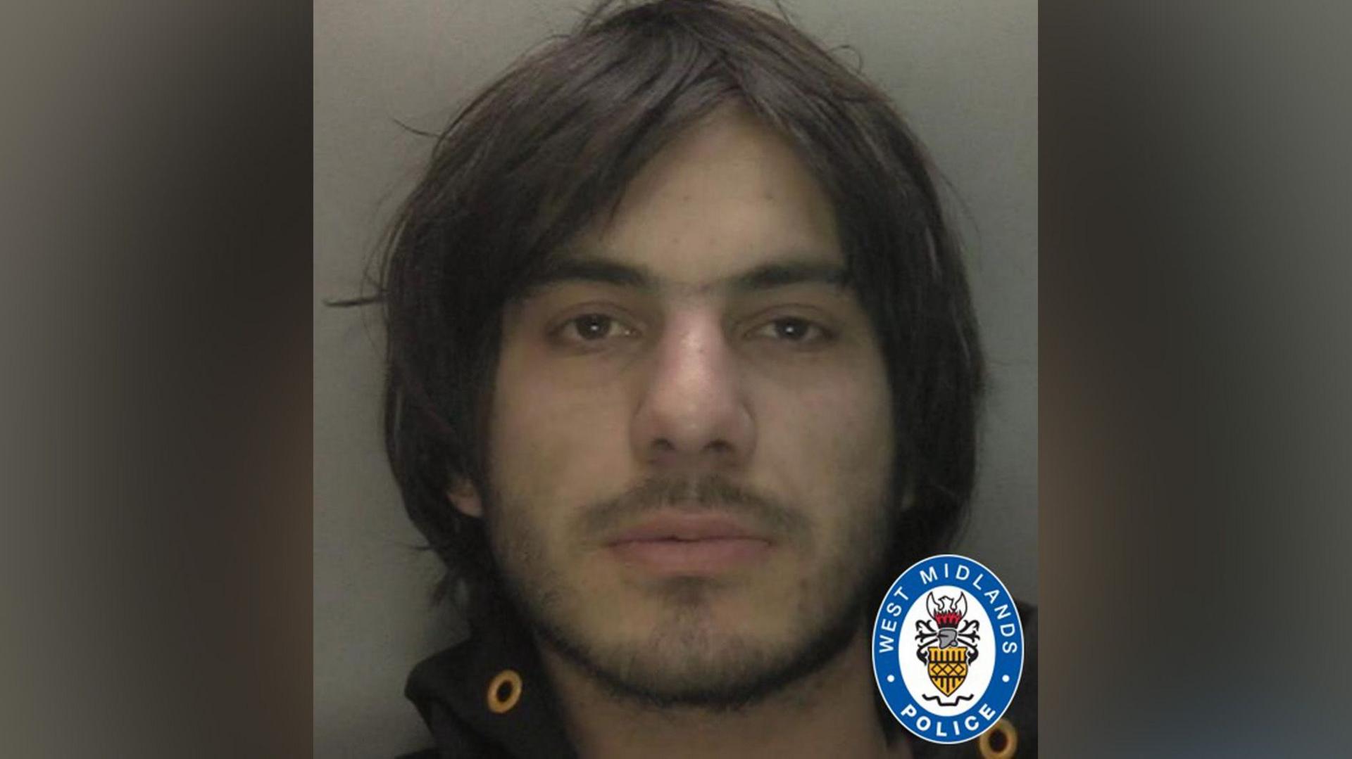 A police mugshot of a man with chin length, dark brown hair, a light beard and brown eyes.