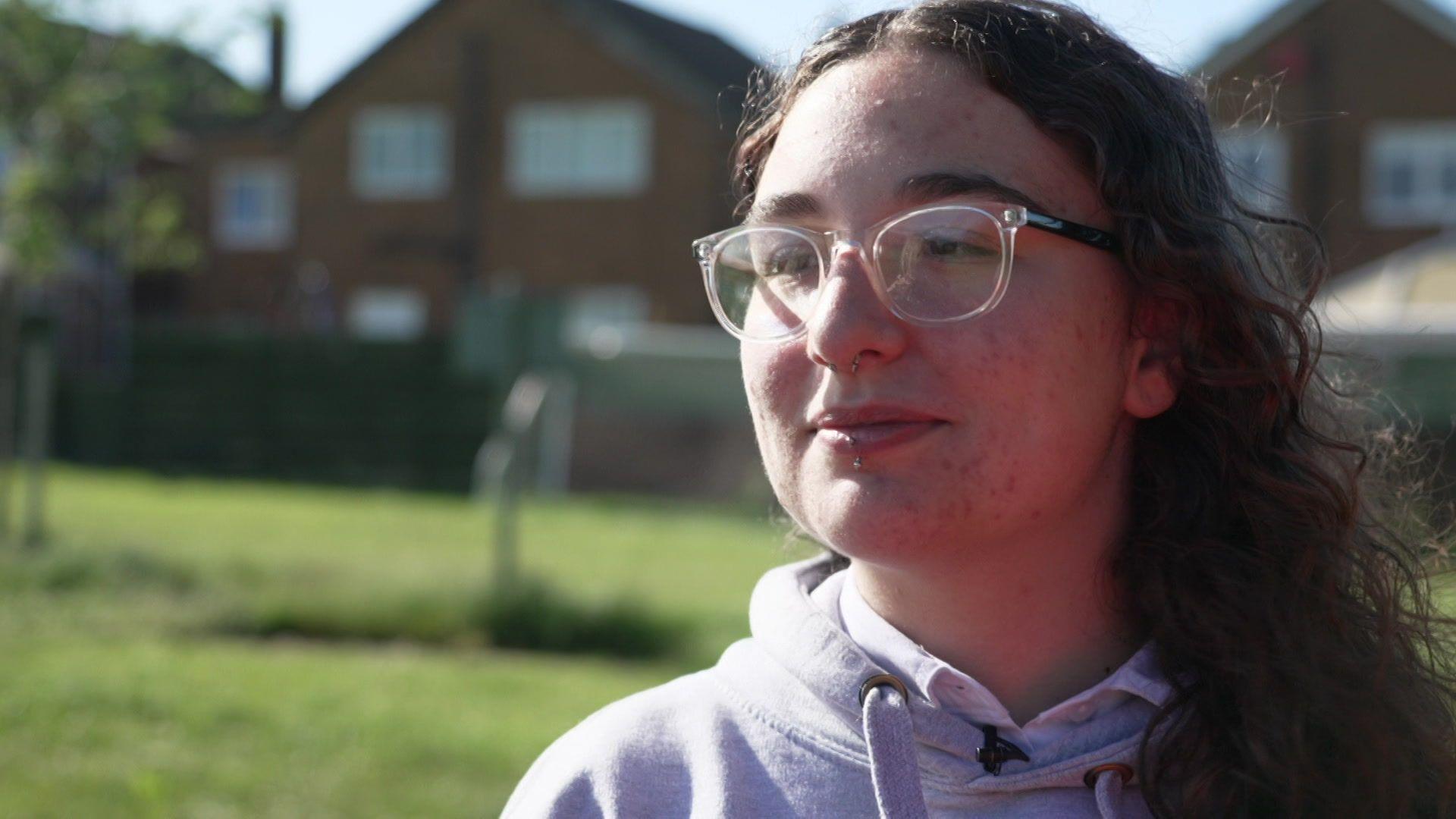 A picture of Mary, she has dark brown curly hair and is wearing glasses. There are houses in the background.