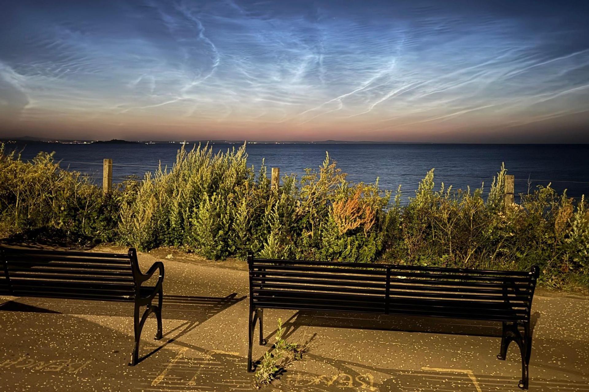 The streaky blue and silver clouds appear on the horizon above the sea. In the foreground are bushes and park benches.