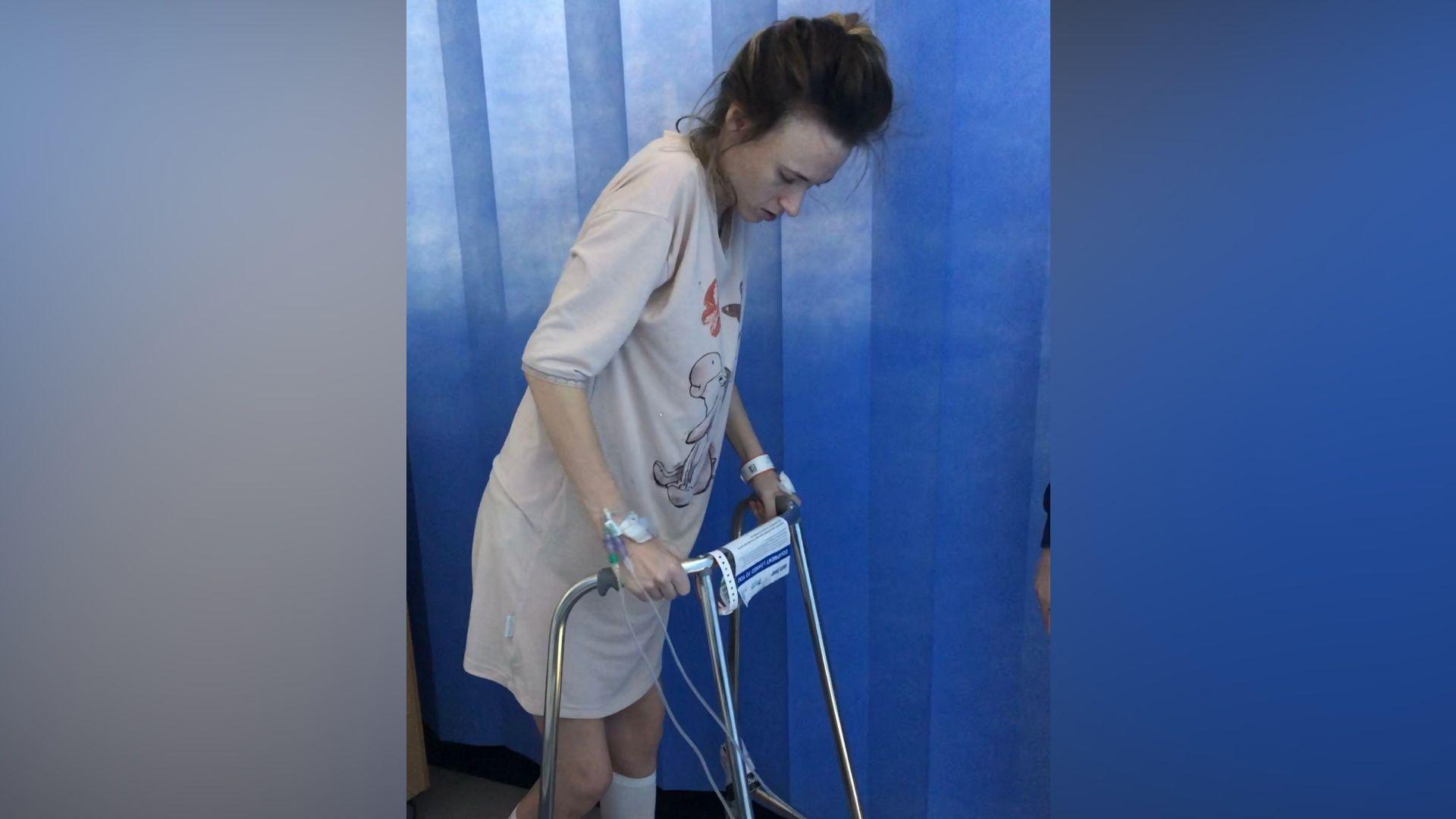 A woman in hospital using a walker. She has dark hair that has been tied up and she is wearing a long pink nightgown. There are various tubes connected to her and hospital machinery. She is walking in front of a blue curtain that separates hospital beds.